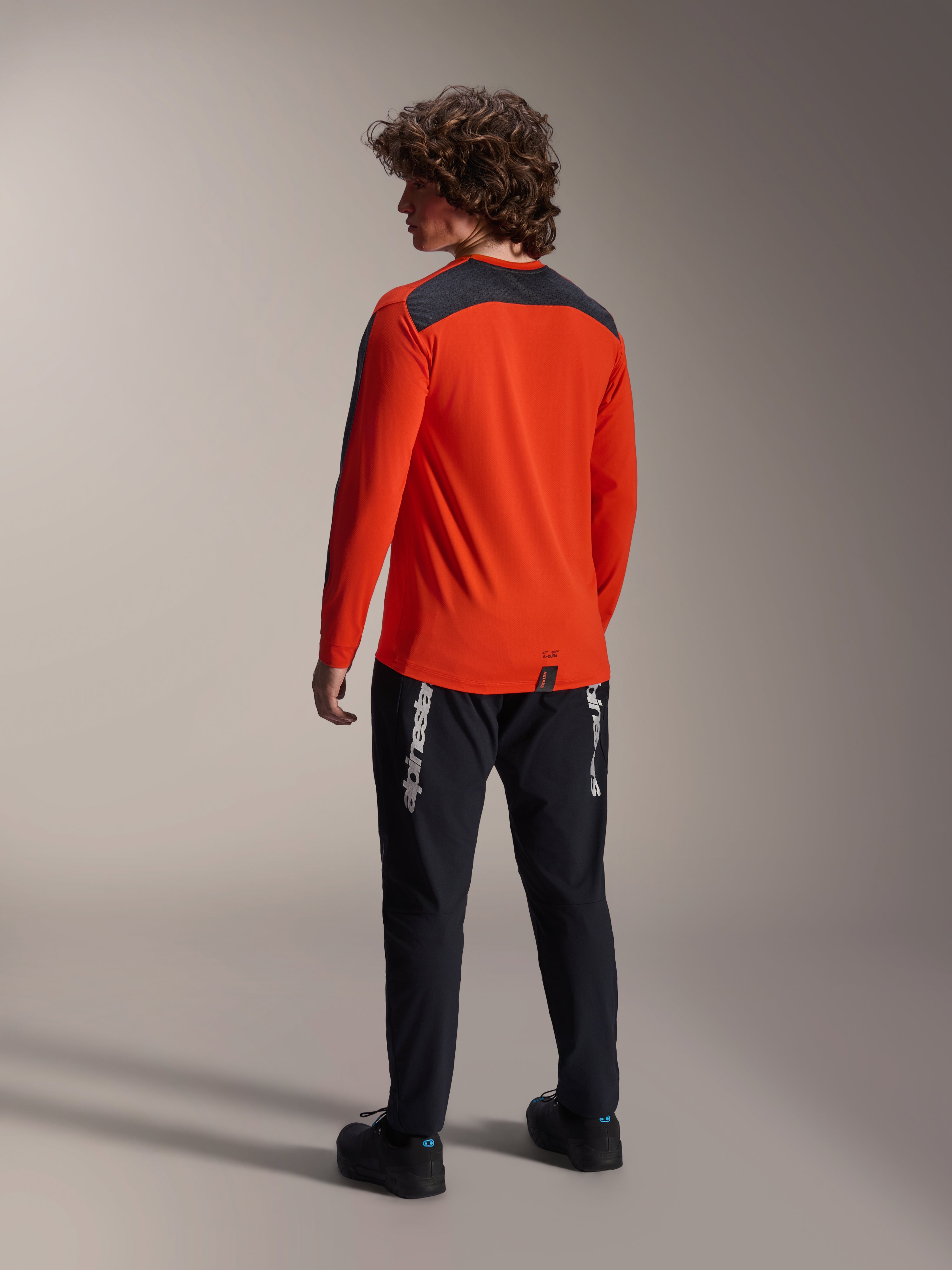 Vue arrière d'un mannequin portant le maillot Alpinestars A-Dura Insert Astar en Spicy Orange, maillot de VTT à manches longues avec empiècement gris foncé aux épaules, associé à un pantalon noir de la marque, équipement de cyclisme tout-terrain.