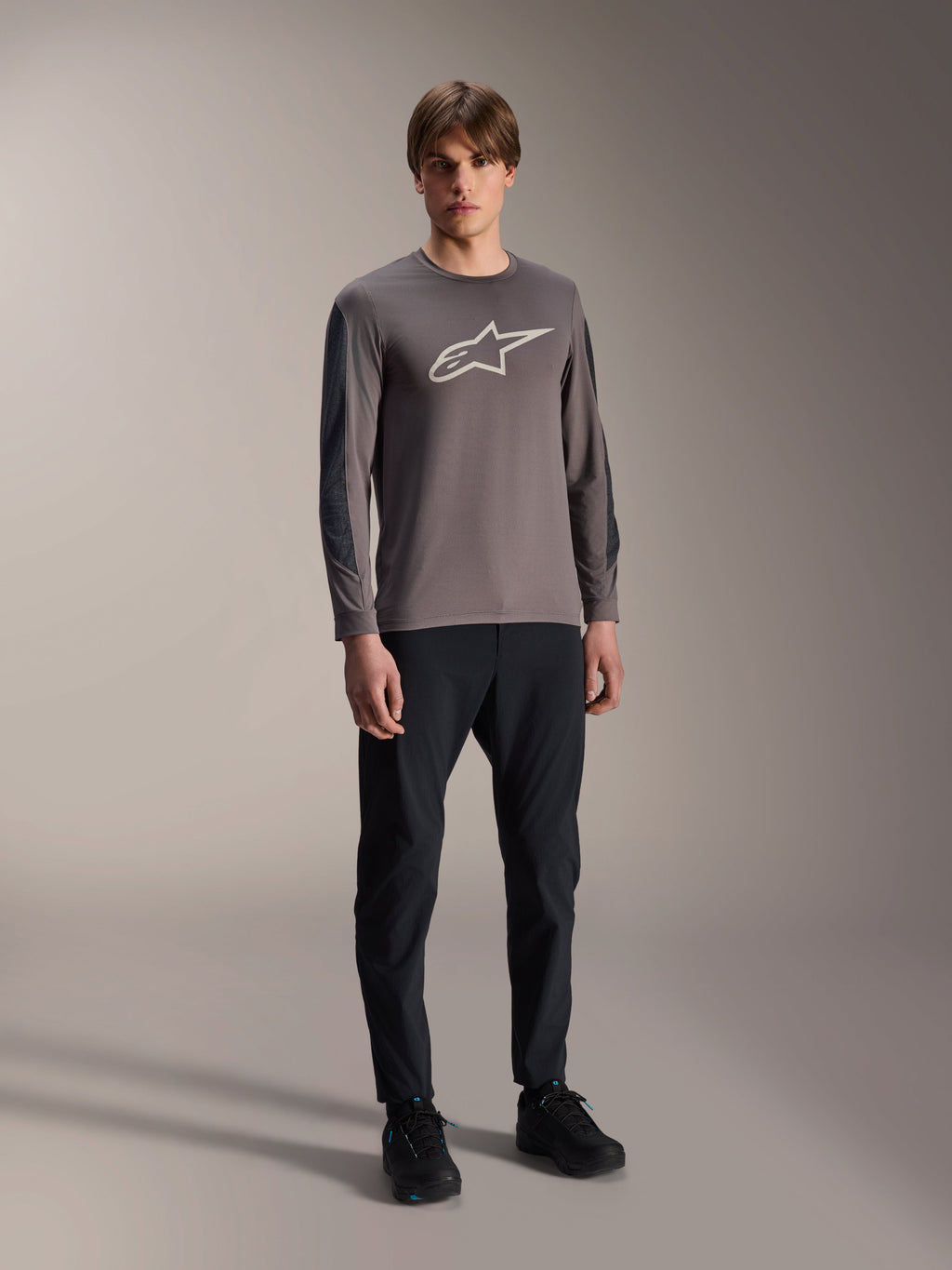 Maillot Alpinestars A-Dura Insert Astar, maillot de VTT à manches longues, gris foncé, avec un grand logo blanc sur la poitrine, porté avec un pantalon technique noir et des chaussures noires, équipement de cyclisme tout-terrain