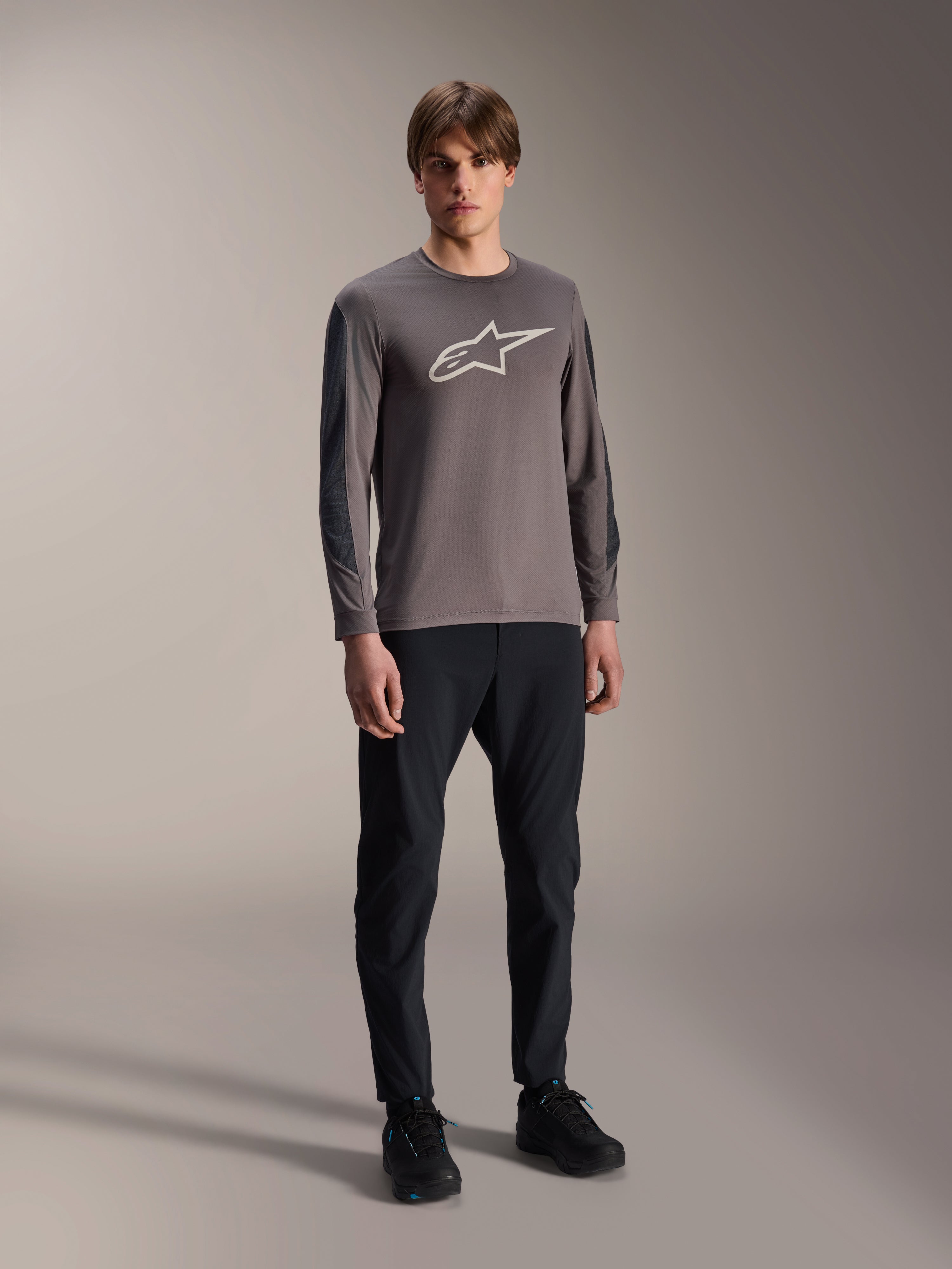 Maillot Alpinestars A-Dura Insert Astar, maillot de VTT à manches longues, gris foncé, avec un grand logo blanc sur la poitrine, porté avec un pantalon technique noir et des chaussures noires, équipement de cyclisme tout-terrain