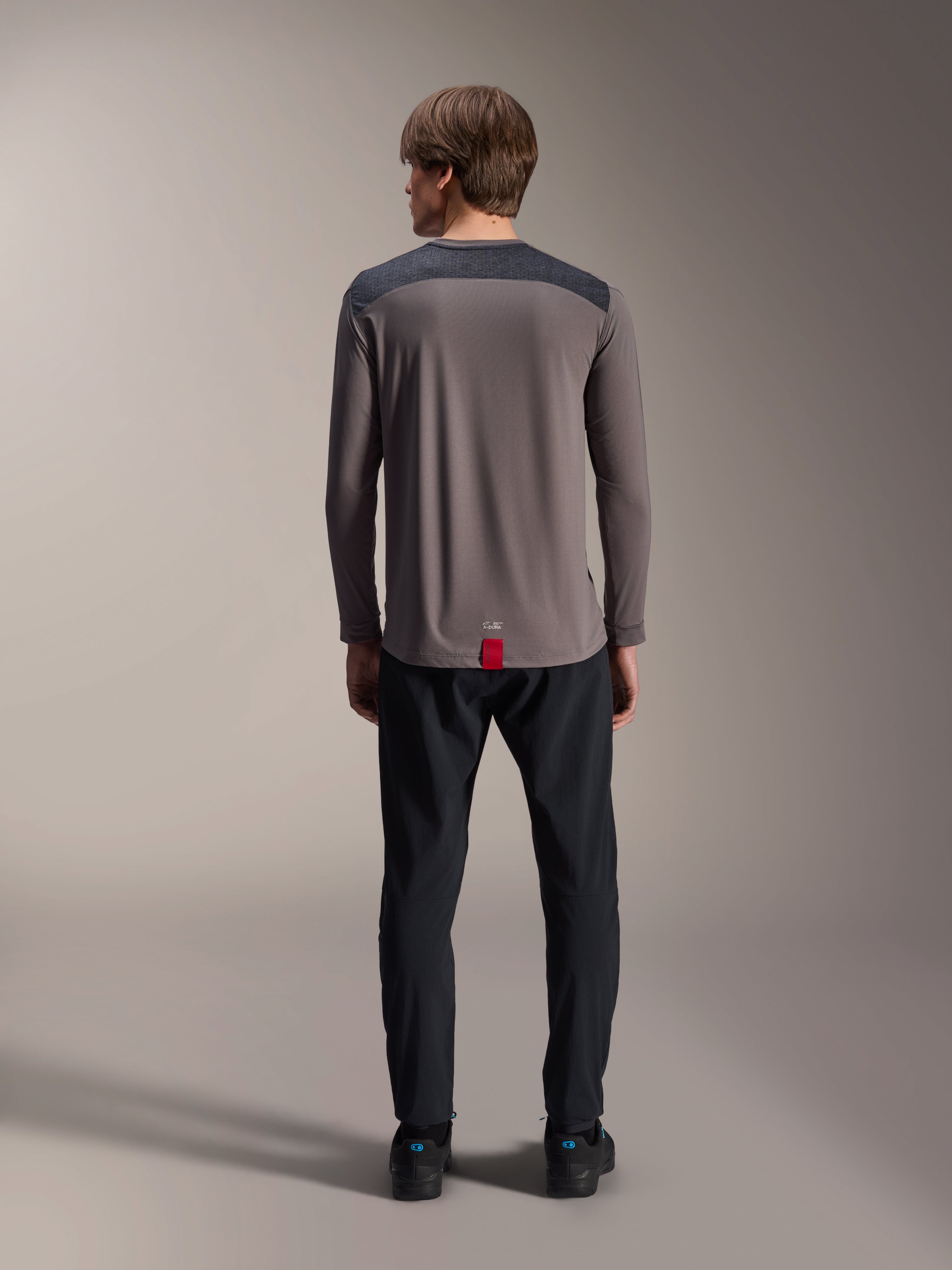 Maillot à manches longues Alpinestars A-Dura Insert Astar en gris foncé, vue de dos, doté d'empiècements anthracite aux épaules et d'une étiquette rouge à l'ourlet, porté avec un pantalon et des chaussures noirs, vêtement de performance pour le VTT