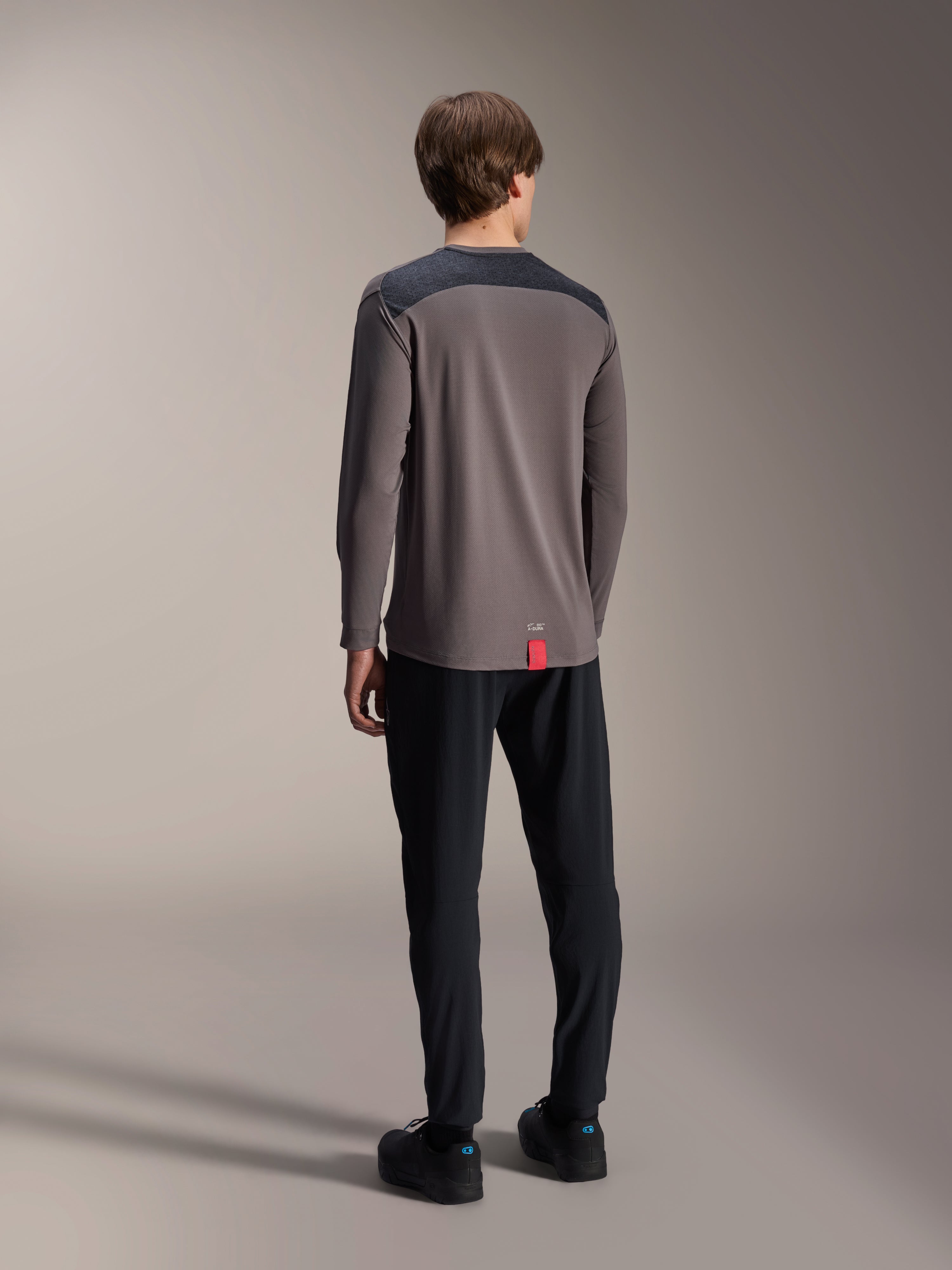 Maillot à manches longues Alpinestars A-Dura Insert Astar, gris foncé, vue de dos sur mannequin, avec empiècements renforcés anthracite aux épaules et une petite étiquette rouge à l'ourlet, porté avec un pantalon et des chaussures noirs, équipement technique de VTT