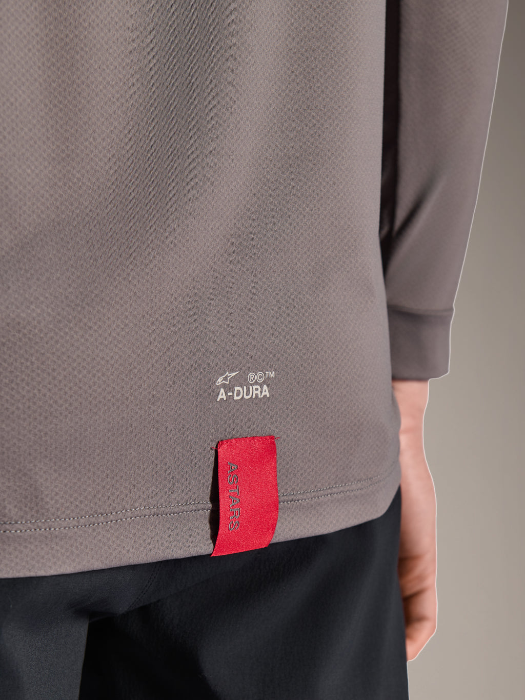 Maillot Alpinestars A-Dura Insert Astar, maillot de VTT à manches longues, gris foncé, gros plan sur le détail de l'ourlet en bas du dos avec marquage A-DURA blanc et étiquette rouge ASTARS emblématique, tissu technique texturé