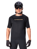 Mountain biker wearing Alpinestars A-Aria Polartec Switch Short Sleeve Jersey in Black, featuring gold-toned chest branding, paired with a black MTB helmet, dark performance sunglasses, and matching gloves, off-road cycling gear.