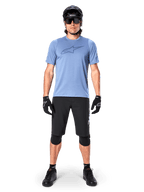 Male mountain bike rider wearing the Alpinestars A-Dura Dri Astar Short Sleeve Jersey in Infinity Blue, featuring a prominent Astars logo on the chest, coordinated with black MTB shorts, black helmet, sunglasses, and protective gear for off-road riding