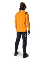 Mountain biker seen from behind wearing the Alpinestars A-Dura Dri Wool Long Sleeve Jersey in Dark Gold, black technical pants with white branding, and a coordinated black and orange helmet, performance-driven mountain bike apparel.