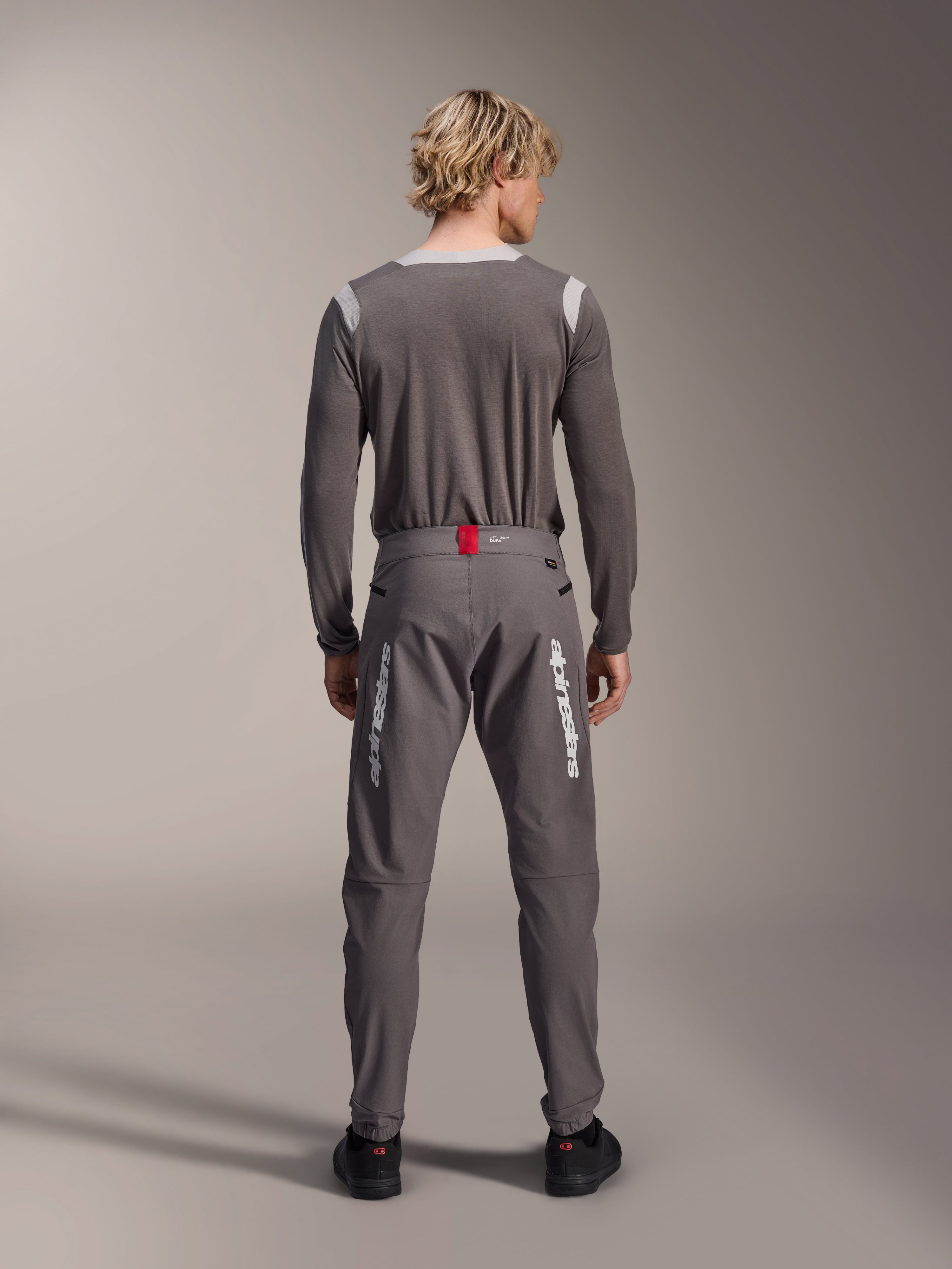 Modèle de dos portant le pantalon Alpinestars A-Dura Elite en gris foncé et un maillot à manches longues assorti, avec des logos Alpinestars blancs sur les jambes, un accent rouge à la taille et des chaussures noires, équipement de VTT professionnel.
