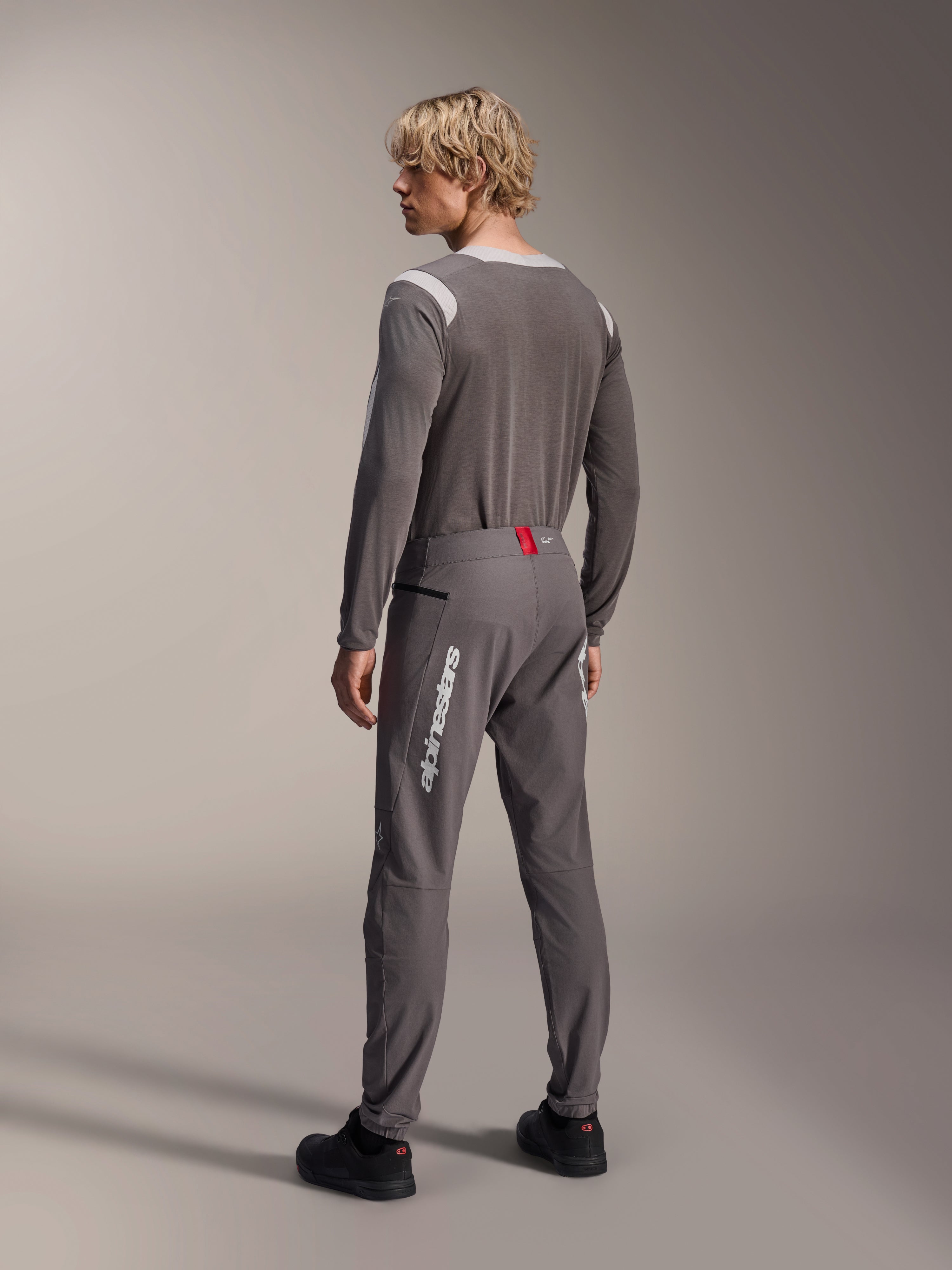 Modèle vu de dos portant un équipement de VTT Alpinestars, maillot à manches longues gris A-Dura Dri Wool, pantalon A-Dura Elite gris foncé avec marquage vertical blanc et détail rouge à la taille, tenue de cyclisme coordonnée pour le VTT