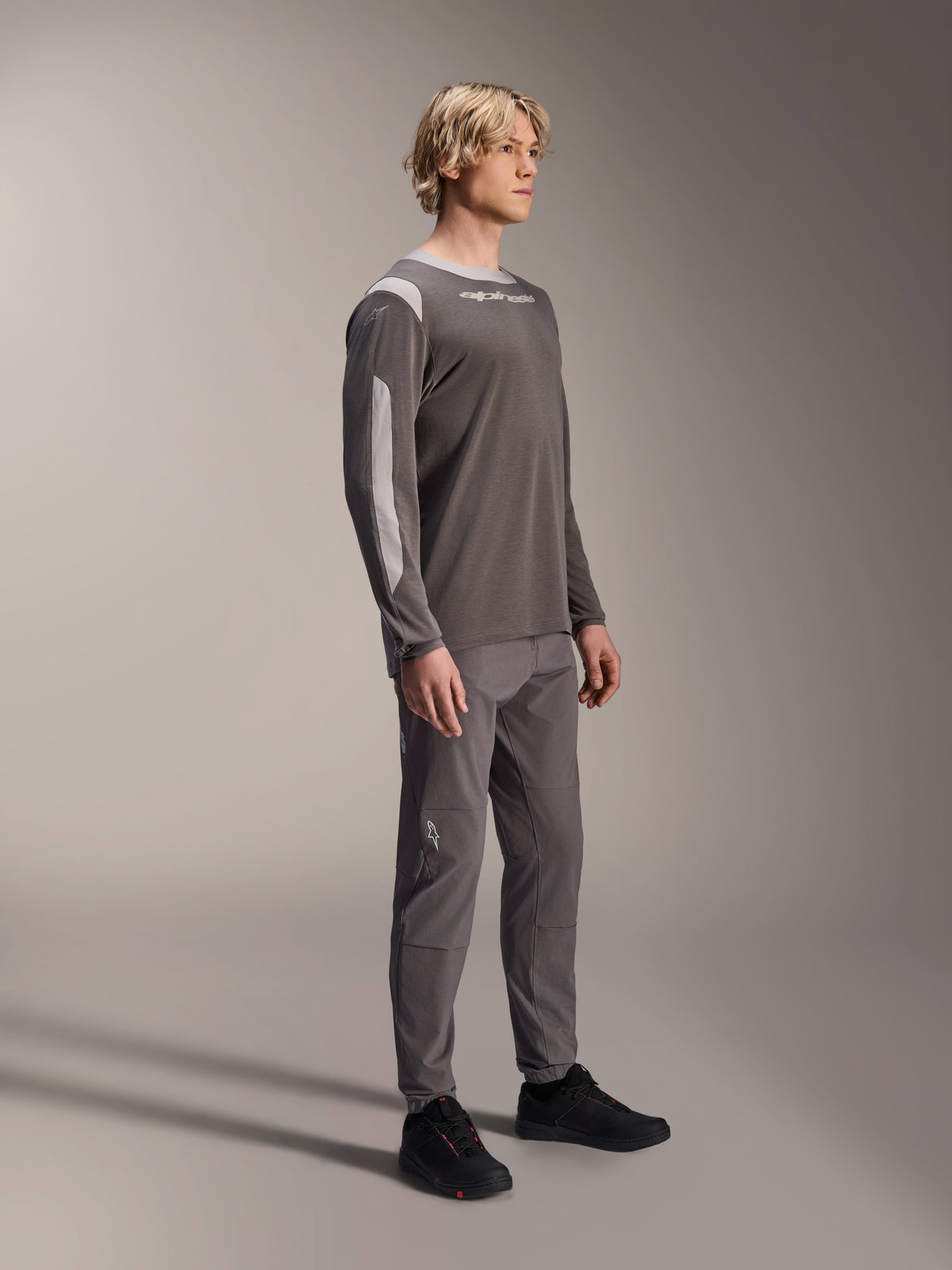 Modèle masculin portant le maillot à manches longues Alpinestars A-Dura Dri Wool en gris foncé, associé à un pantalon gris assorti et des chaussures noires, avec des panneaux en maille gris clair sur les épaules et les manches, équipement technique de VTT pour le cyclisme tout-terrain