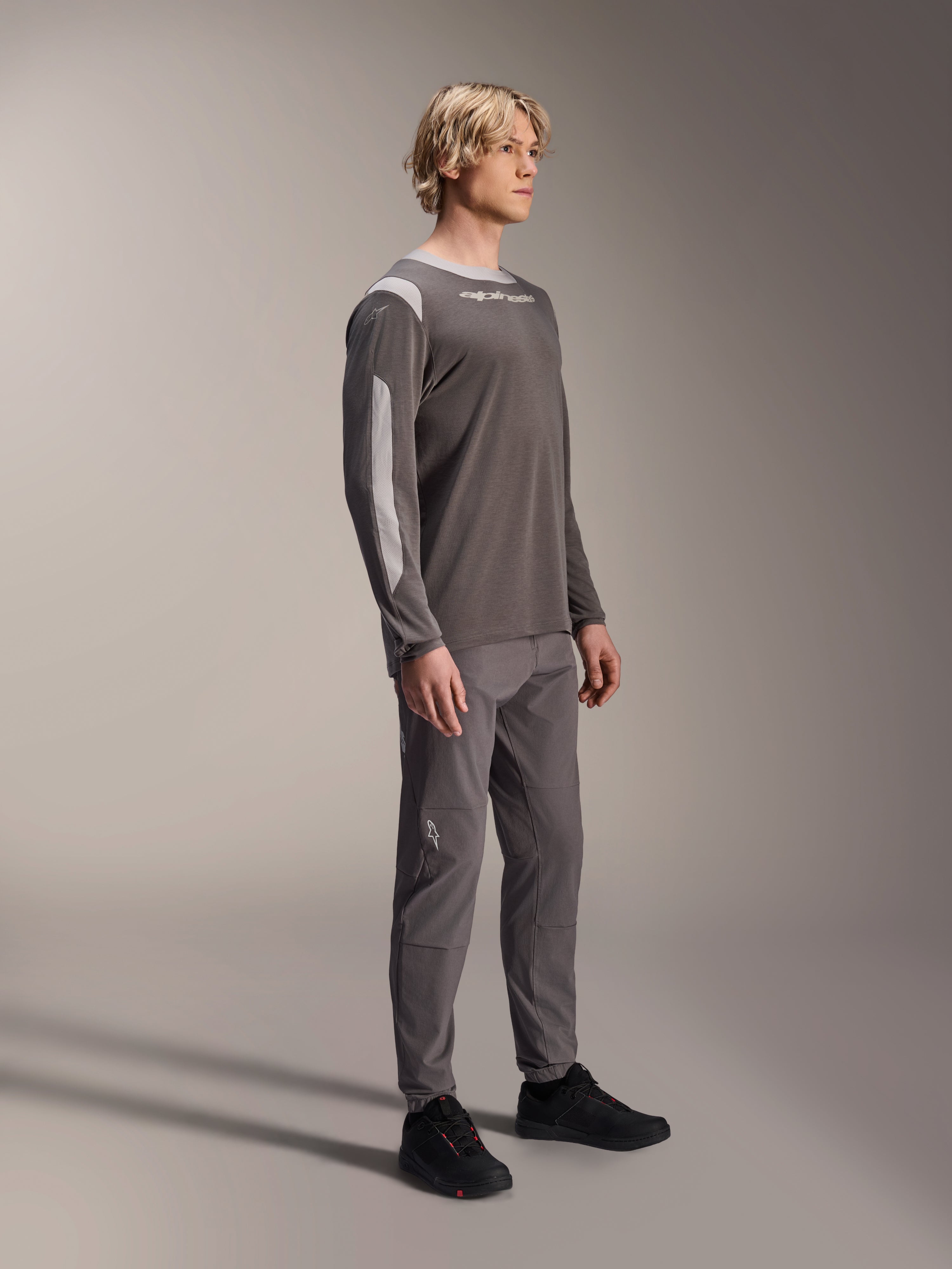 Modèle masculin portant le maillot à manches longues Alpinestars A-Dura Dri Wool en gris foncé, associé à un pantalon gris assorti et des chaussures noires, avec des panneaux en maille gris clair sur les épaules et les manches, équipement technique de VTT pour le cyclisme tout-terrain