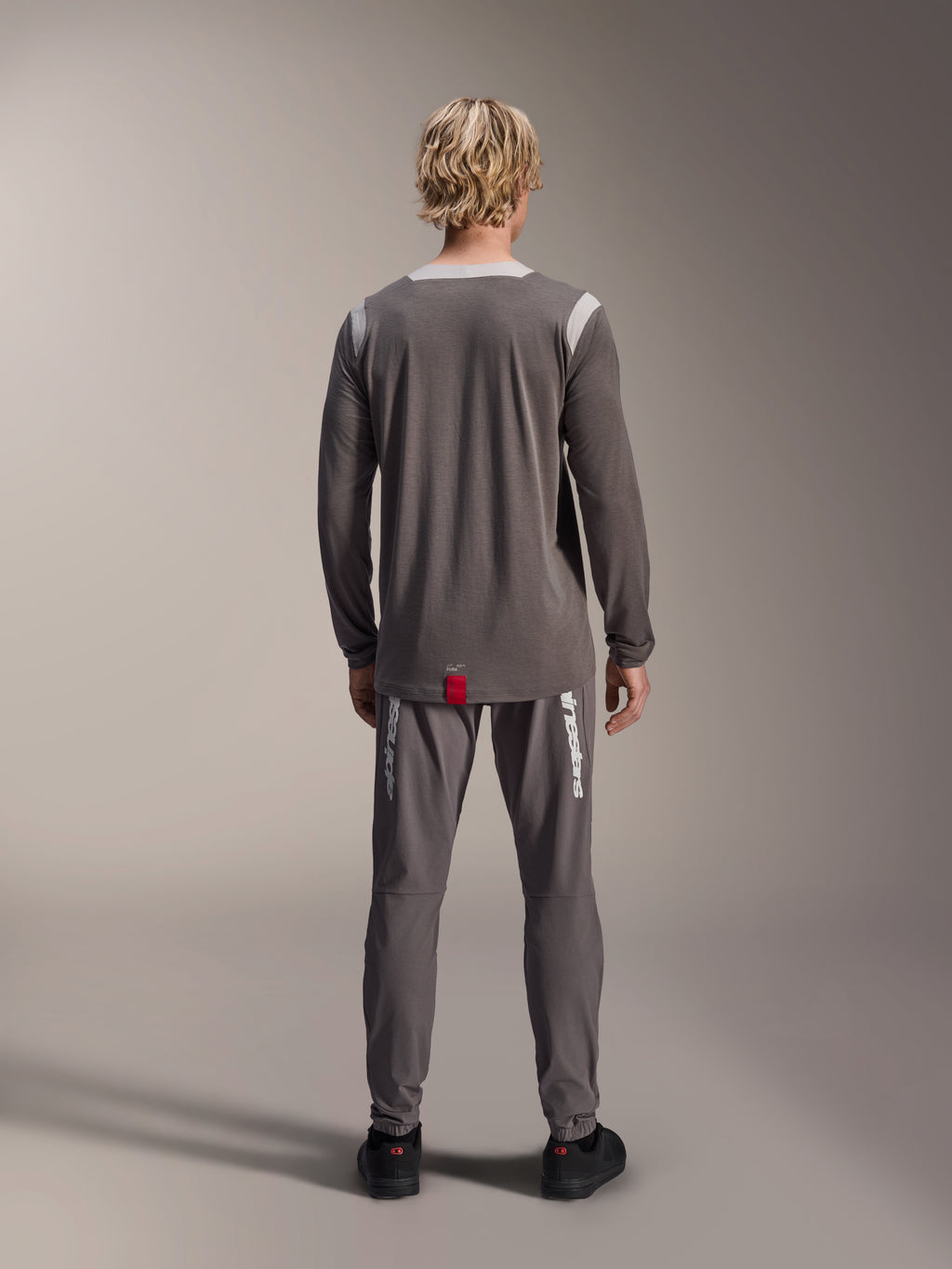 Maillot à manches longues Alpinestars A-Dura Dri Wool, gris foncé, vue de dos sur modèle, avec panneaux en maille gris clair sur les épaules et le col, pan arrière allongé avec une petite étiquette rouge, porté avec un pantalon de cyclisme gris assorti