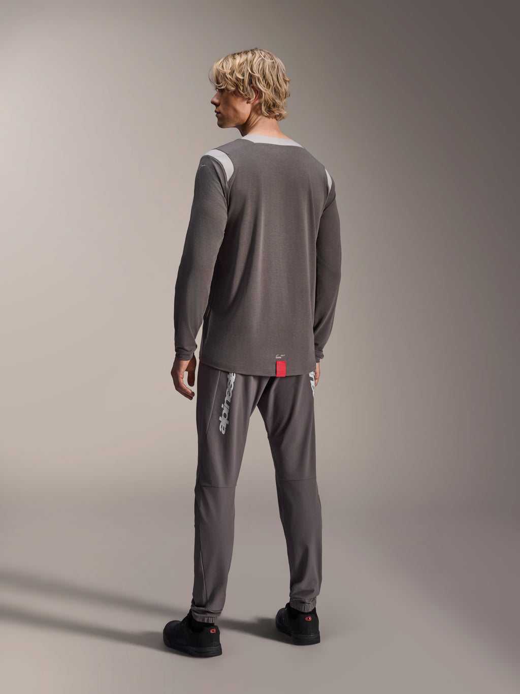 Mannequin homme portant le maillot à manches longues Alpinestars A-Dura Dri Wool, gris foncé avec empiècements en mesh gris clair aux épaules, associé à un pantalon de cyclisme gris foncé assorti, vue de dos montrant le pan arrière allongé, tenue de VTT