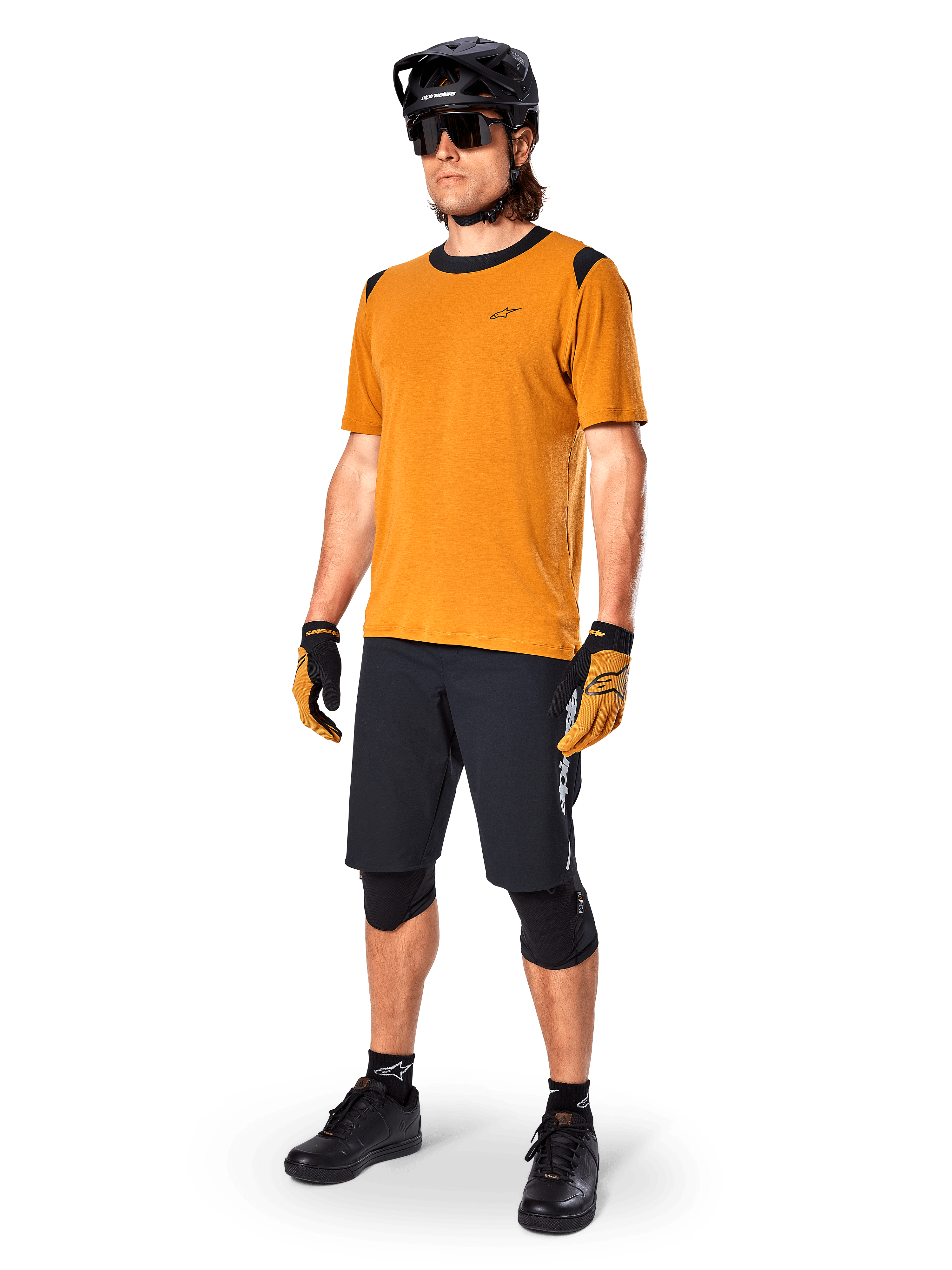 Male model wearing Alpinestars A-Dura Dri Wool Jersey - Short Sleeve in Dark Gold, mountain bike jersey with black shoulder and collar accents, paired with black MTB shorts, black helmet, and sunglasses, technical off-road cycling gear