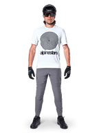 Mountain biker wearing Alpinestars A-Dura Oscar Jersey, Short Sleeve, White Dove with large black spiral logo, paired with grey pants, black helmet, sunglasses, and black gloves, performance MTB trail riding gear