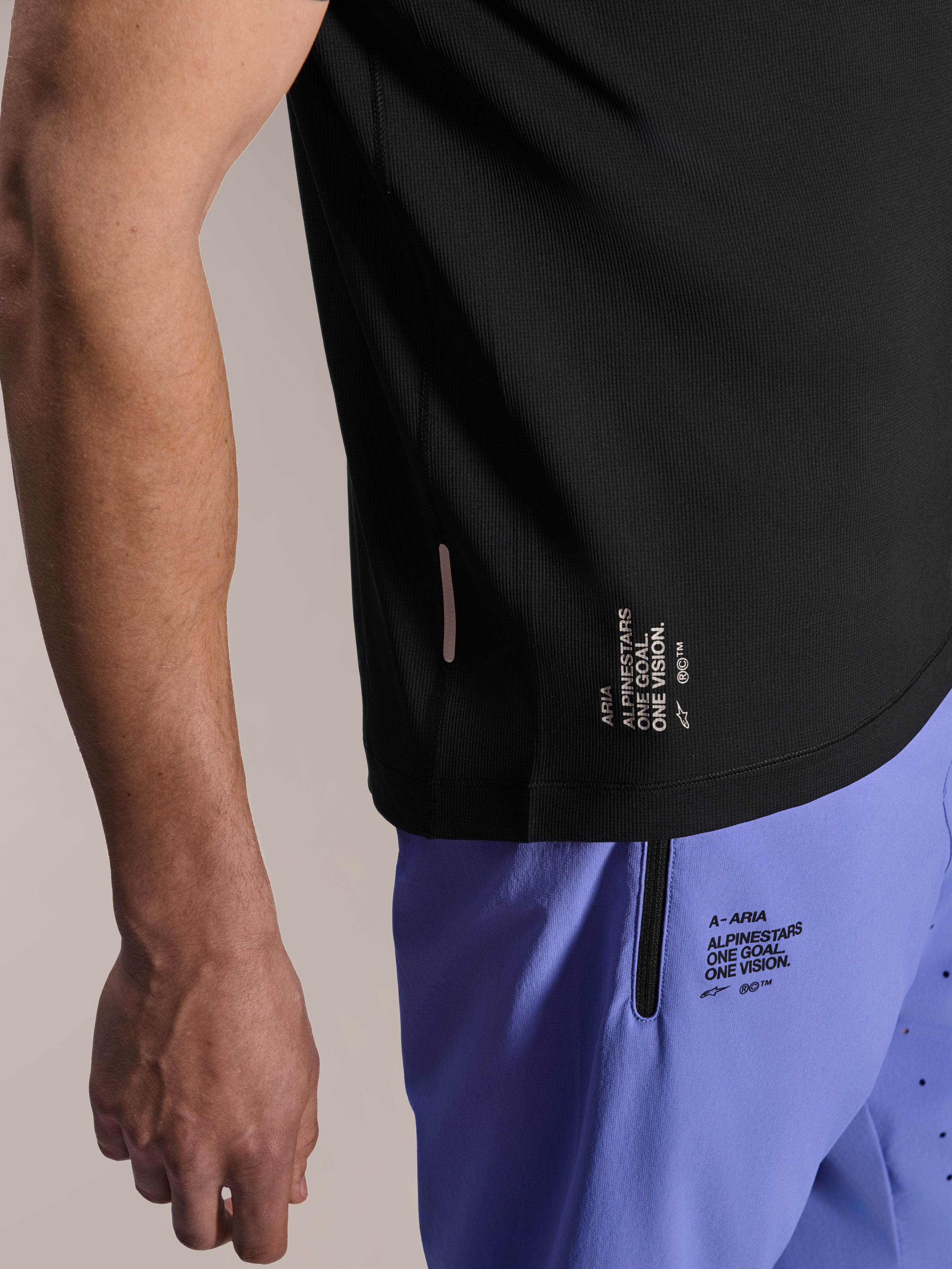 Alpinestars A-Aria Switch Short Sleeve Jersey, Black MTB jersey, close-up of side hem with white 'ONE GOAL. ONE VISION.' branding, worn with light purple cycling shorts featuring matching logos
