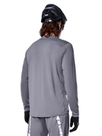 Alpinestars A-Dura Rocker Long Sleeve Jersey in Dark Gray, back view of mountain bike rider, paired with matching gray shorts featuring white branding, black MTB helmet, and black gloves, professional off-road cycling gear.