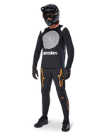 MTB rider wearing Alpinestars A-Supra Oscar Long Sleeve Jersey in Black, featuring the white vintage Oscar spiral graphic, black pants with orange star logos, black full-face helmet with goggles, and black gloves, professional mountain biking gear