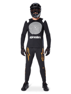 Male model wearing complete mountain bike gear, Alpinestars A-Supra Oscar Long Sleeve Jersey in black with white spiral chest graphic, black pants with orange star logos, black full-face helmet and goggles, black gloves and shoes, coordinated outfit for MTB gravity riding