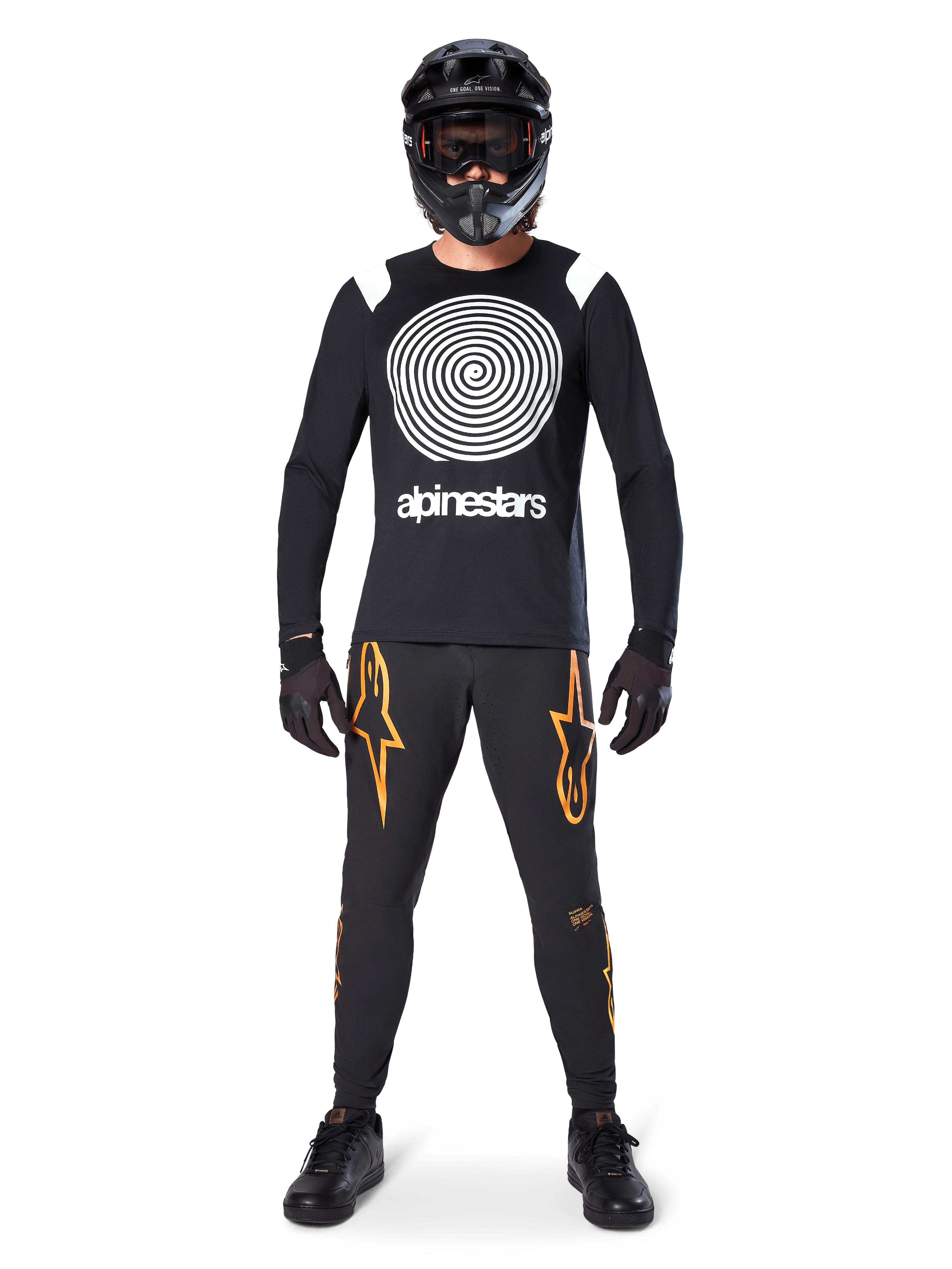 Male model wearing complete mountain bike gear, Alpinestars A-Supra Oscar Long Sleeve Jersey in black with white spiral chest graphic, black pants with orange star logos, black full-face helmet and goggles, black gloves and shoes, coordinated outfit for MTB gravity riding