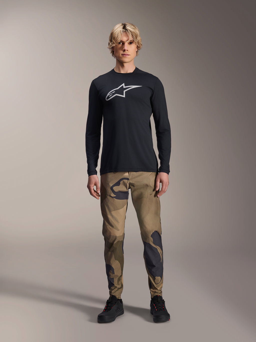 Maillot à manches longues Alpinestars A-Dura Astar, noir, arborant un grand logo Astars blanc sur la poitrine, porté avec un pantalon camouflage et des chaussures noires, tenue de performance décontractée pour le VTT