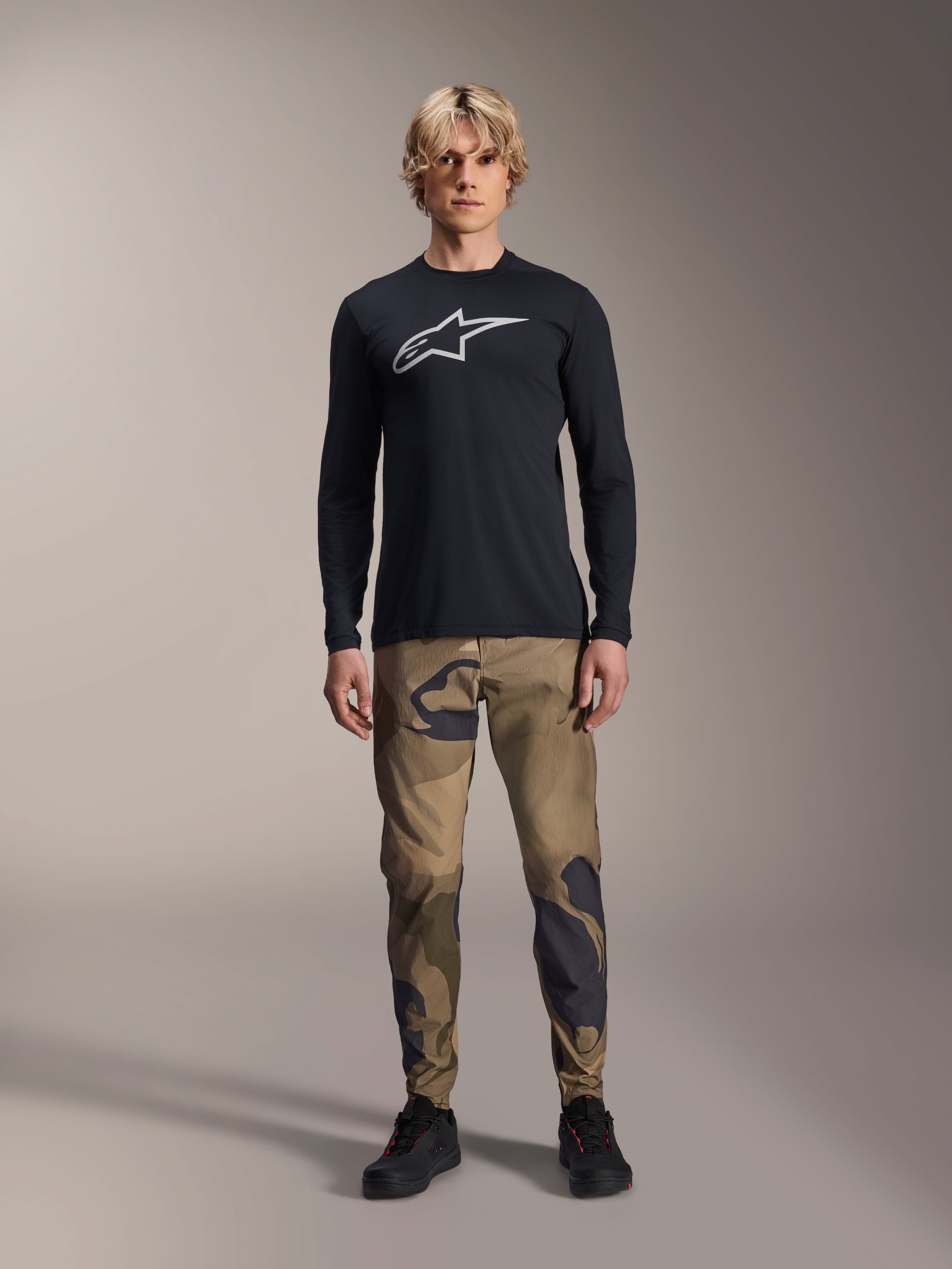 Maillot à manches longues Alpinestars A-Dura Astar, noir, arborant un grand logo Astars blanc sur la poitrine, porté avec un pantalon camouflage et des chaussures noires, tenue de performance décontractée pour le VTT