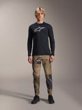 Alpinestars A-Dura Camo Pant, pantalon de cyclisme VTT, camouflage marron et vert, porté avec un maillot noir à manches longues et des chaussures noires, style VTT sportif avec tissu extensible durable