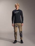 Alpinestars A-Dura Camo Pant, pantalon de cyclisme VTT, camouflage marron et vert, porté avec un maillot noir à manches longues et des chaussures noires, style VTT sportif avec tissu extensible durable