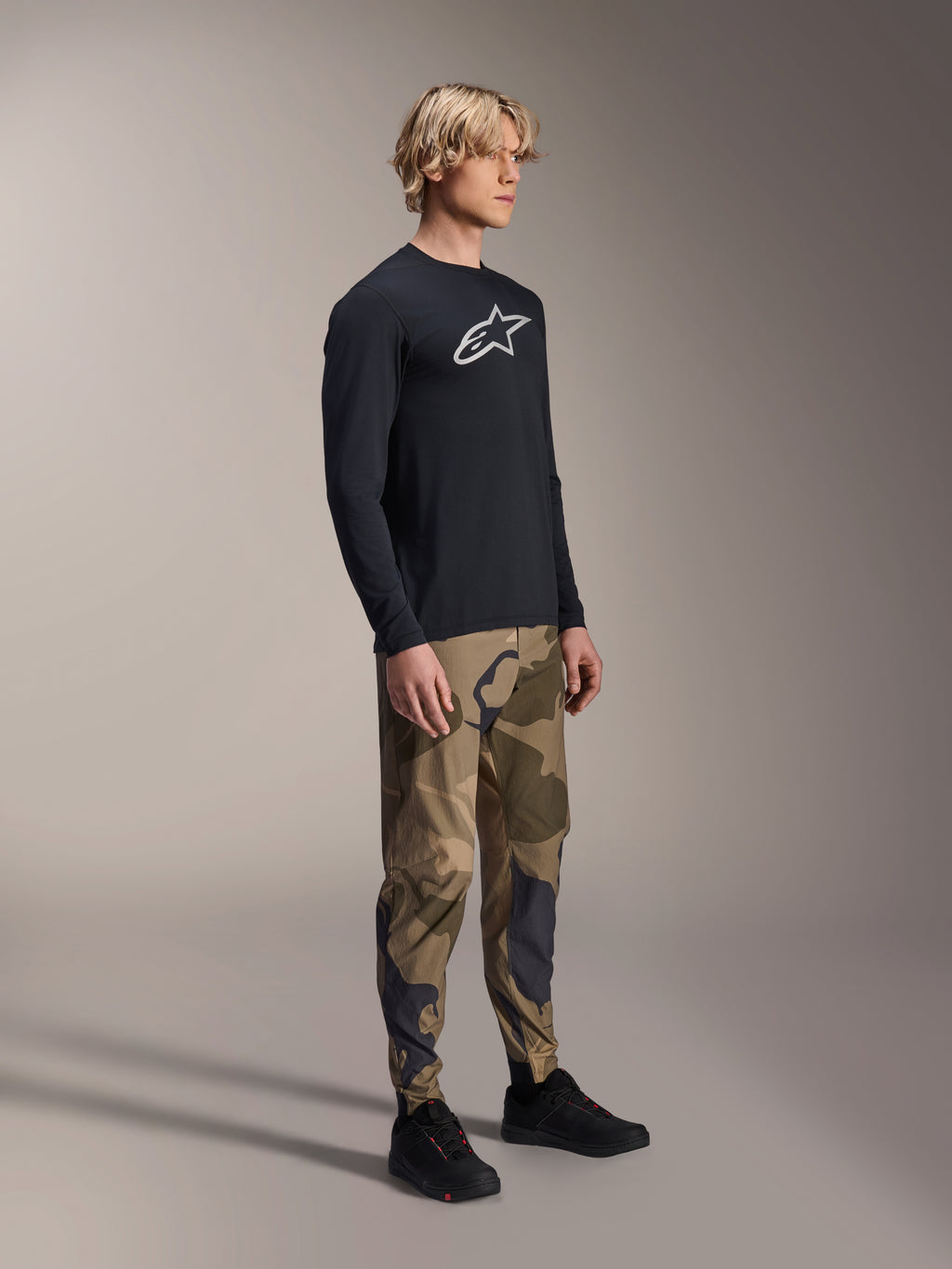 Maillot à manches longues Alpinestars A-Dura Astar en noir, porté par un mannequin avec un pantalon camouflage et des baskets noires, logo Astars blanc sur la poitrine, vêtement de performance VTT décontracté pour le trail.