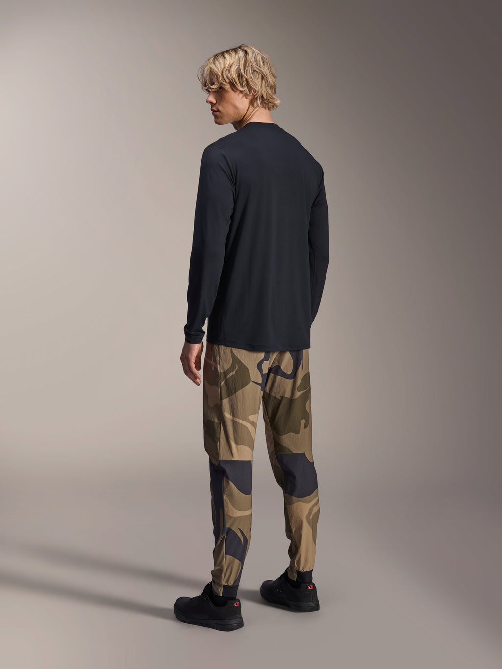 Alpinestars A-Dura Camo Pant, pantalon de VTT, camouflage brun, porté avec un maillot à manches longues noir et des baskets noires, vue arrière montrant des jambes fuselées et des chevilles élastiquées, vêtement de performance pour le cyclisme tout-terrain