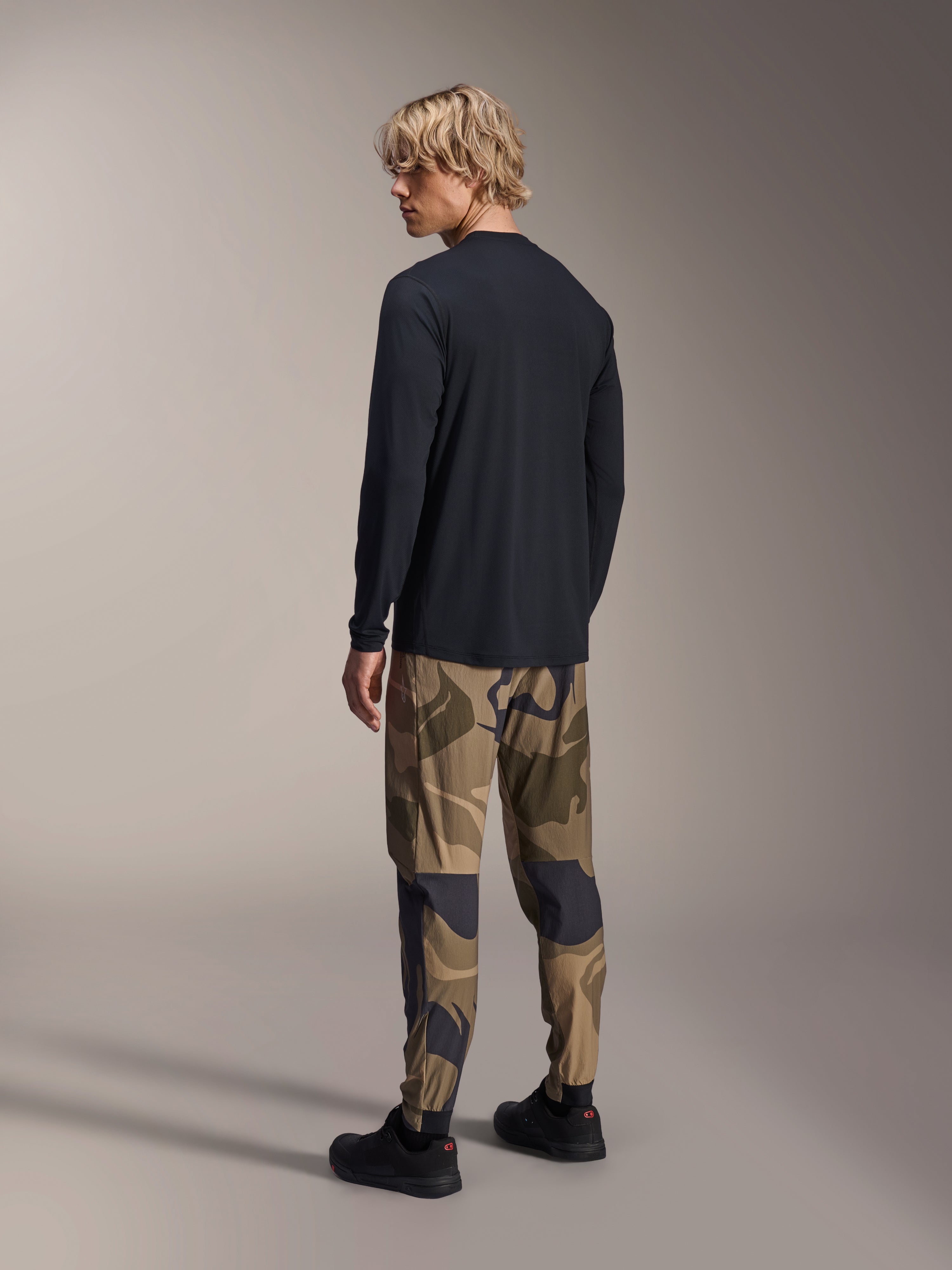 Alpinestars A-Dura Camo Pant, pantalon de VTT, camouflage brun, porté avec un maillot à manches longues noir et des baskets noires, vue arrière montrant des jambes fuselées et des chevilles élastiquées, vêtement de performance pour le cyclisme tout-terrain
