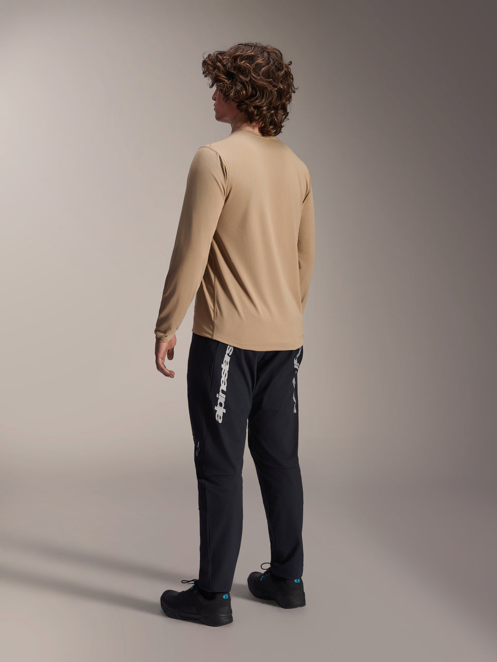 Maillot à manches longues Alpinestars A-Dura Astar en brun clair, vue de dos d'un modèle masculin mettant en valeur la coupe à pan arrière allongé, associé à un pantalon et des chaussures noirs, équipement de performance décontracté pour le VTT