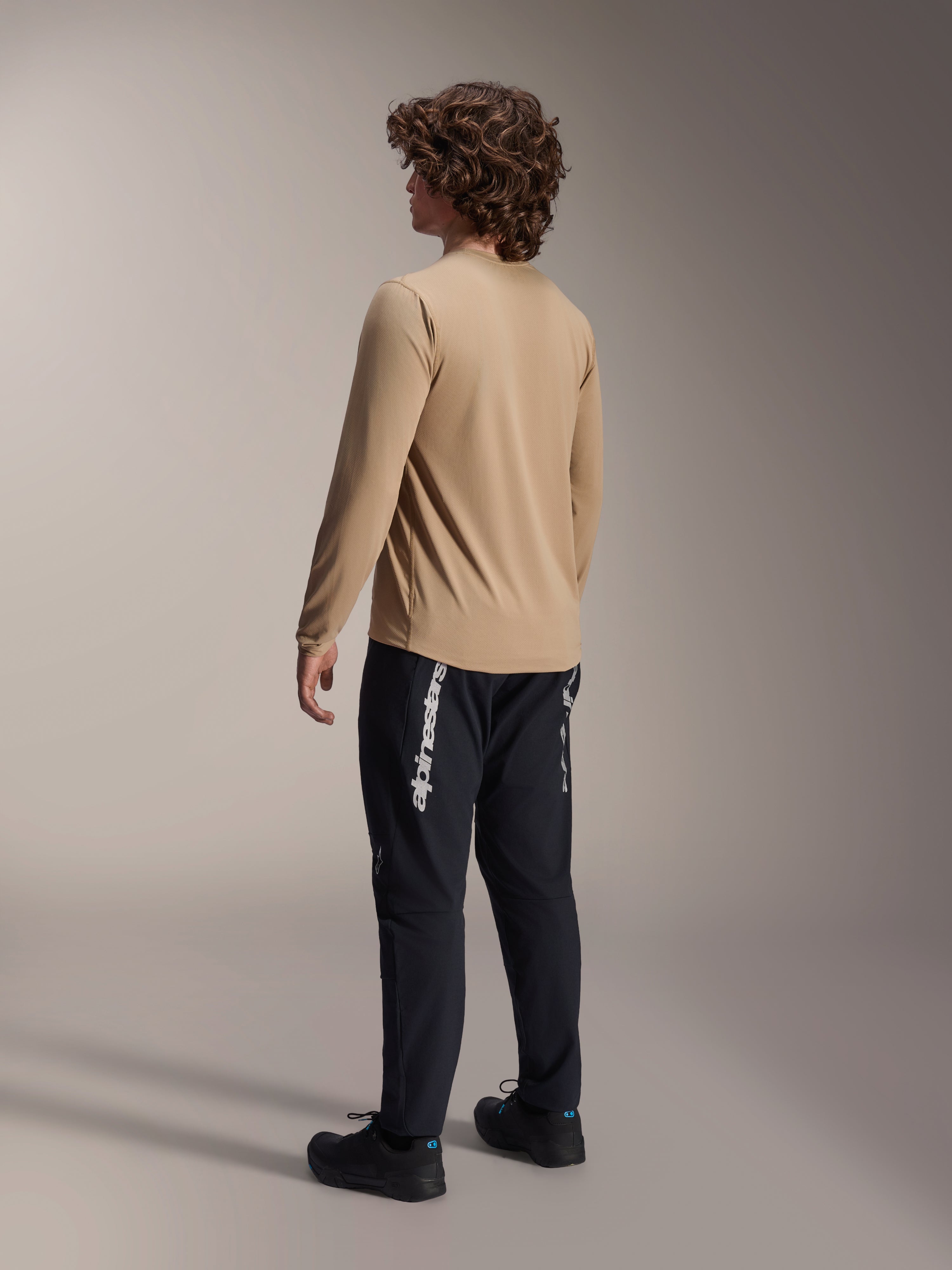 Maillot à manches longues Alpinestars A-Dura Astar en brun clair, vue de dos d'un modèle masculin mettant en valeur la coupe à pan arrière allongé, associé à un pantalon et des chaussures noirs, équipement de performance décontracté pour le VTT