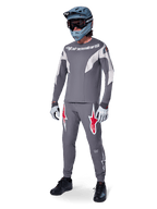 Pilote de VTT portant un maillot Alpinestars A-Supra Race à manches longues, gris foncé avec des accents gris clair, pantalon gris foncé assorti avec logo étoile rouge, casque intégral gris avec lunettes, gants gris clair, tenue coordonnée pour la course de VTT