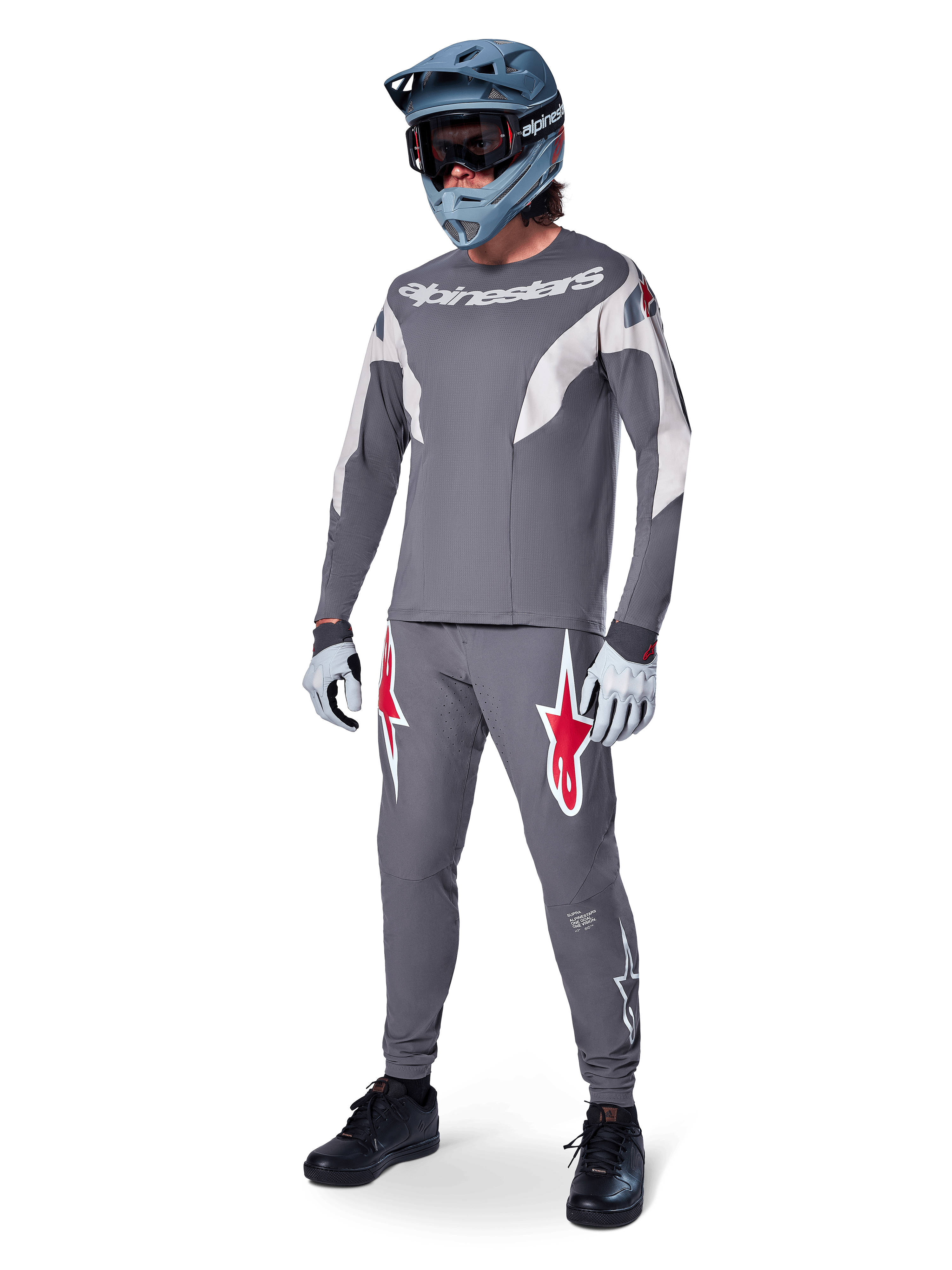Pilote de VTT portant un maillot Alpinestars A-Supra Race à manches longues, gris foncé avec des accents gris clair, pantalon gris foncé assorti avec logo étoile rouge, casque intégral gris avec lunettes, gants gris clair, tenue coordonnée pour la course de VTT