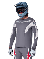 Maillot Alpinestars A-Supra Race à manches longues, gris foncé avec accents blancs et rouges, graphismes de style course, porté avec un pantalon gris assorti, un casque intégral bleu et des gants blancs, équipement professionnel de VTT et de descente