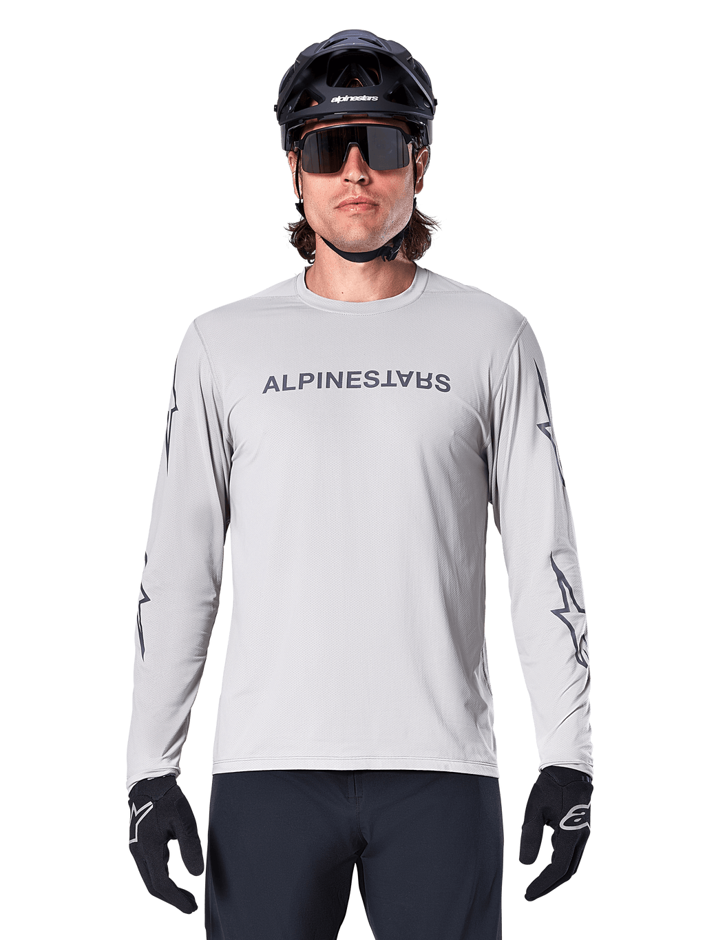 Maillot à manches longues Alpinestars A-Dura Switch, gris clair, avec logo textuel gris foncé sur la poitrine et logos étoiles sur les manches, porté par un cycliste avec un casque de VTT noir et des gants, vêtement technique performant pour le trail