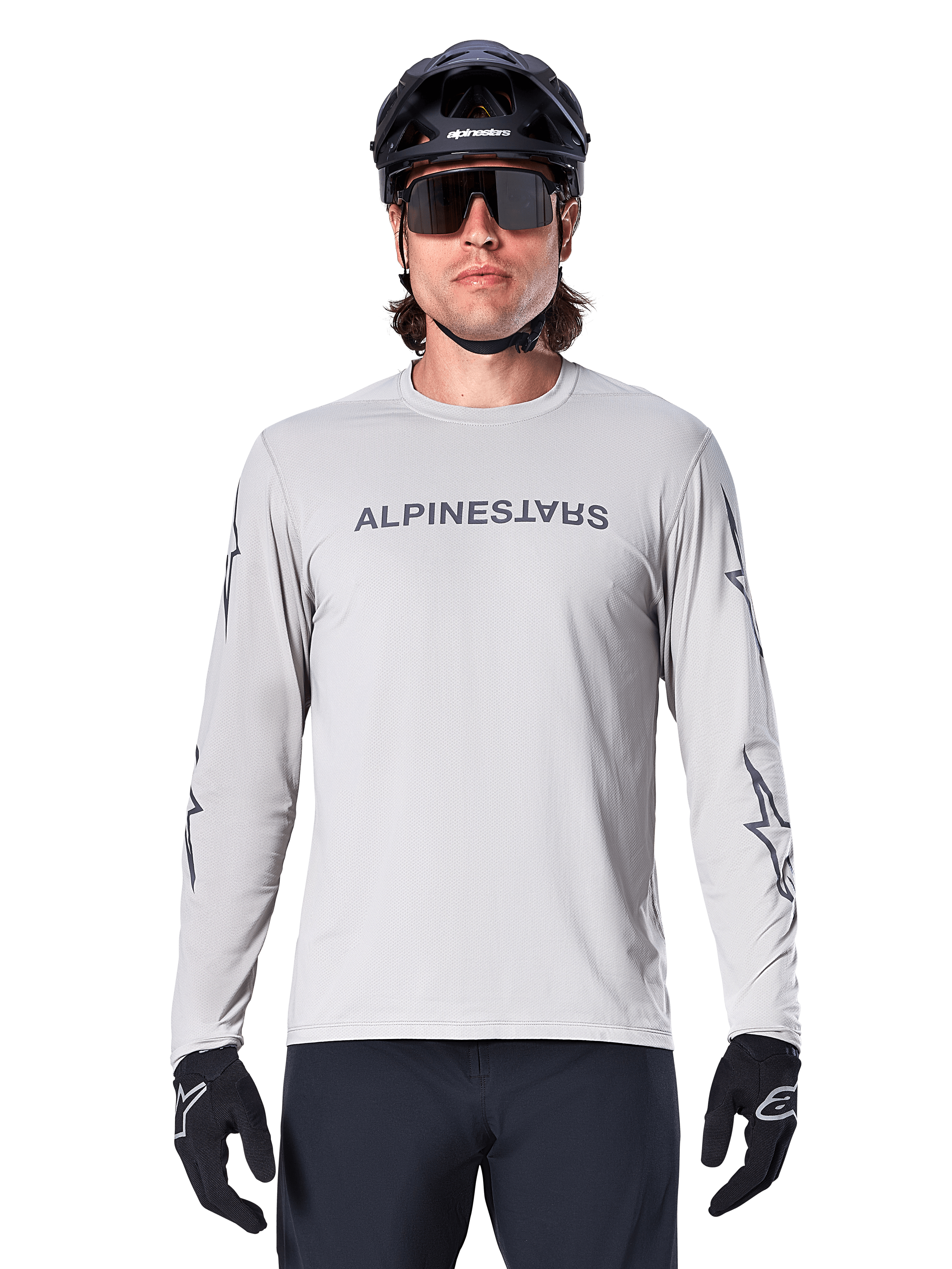 Maillot à manches longues Alpinestars A-Dura Switch, gris clair, avec logo textuel gris foncé sur la poitrine et logos étoiles sur les manches, porté par un cycliste avec un casque de VTT noir et des gants, vêtement technique performant pour le trail