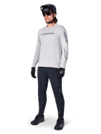 Maillot à manches longues Alpinestars A-Dura Switch, gris clair, porté par un cycliste avec un pantalon de VTT noir et un casque, arborant un logo lettrage noir sur la poitrine et des graphismes d'étoiles sur les manches, équipement de cyclisme tout-terrain haute performance