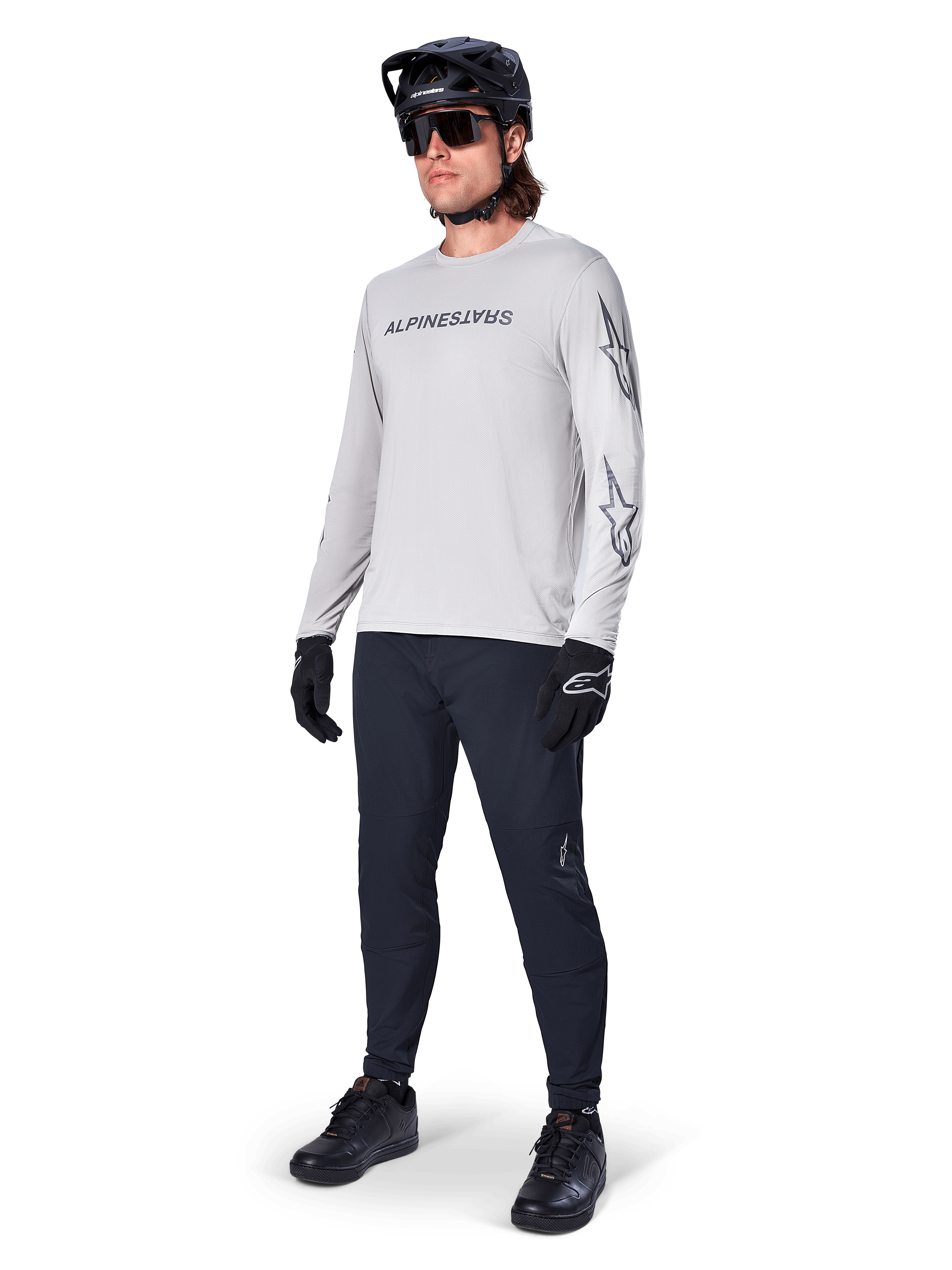 Maillot à manches longues Alpinestars A-Dura Switch, gris clair, porté par un cycliste avec un pantalon de VTT noir et un casque, arborant un logo lettrage noir sur la poitrine et des graphismes d'étoiles sur les manches, équipement de cyclisme tout-terrain haute performance