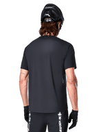 Maillot à manches courtes Alpinestars A-Dura Switch, noir, vue arrière d'un mannequin portant un casque de cyclisme noir et un short de VTT, mettant en avant la coupe régulière et le pan arrière allongé conçu pour le VTT.