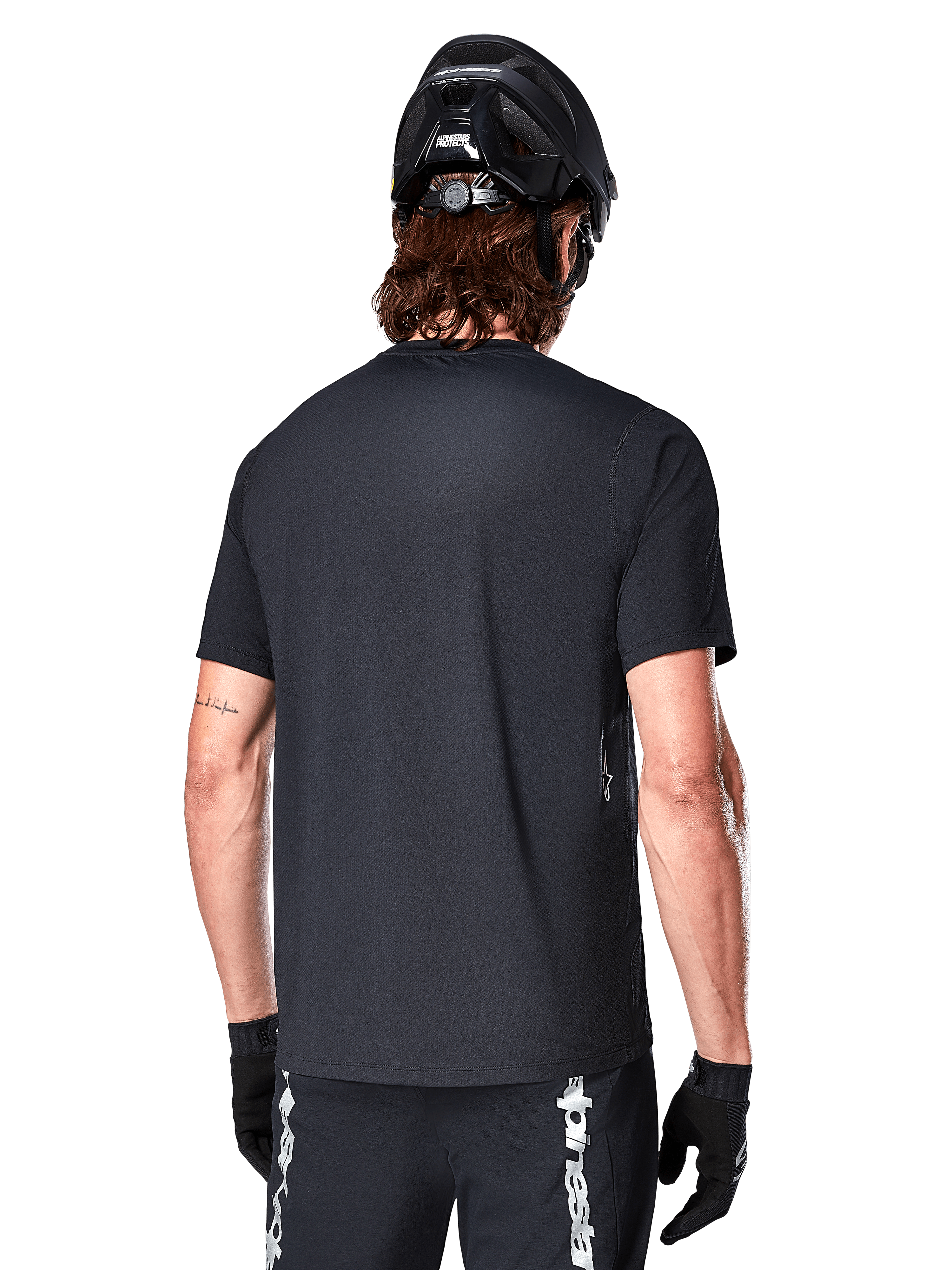 Maillot à manches courtes Alpinestars A-Dura Switch, noir, vue arrière d'un mannequin portant un casque de cyclisme noir et un short de VTT, mettant en avant la coupe régulière et le pan arrière allongé conçu pour le VTT.