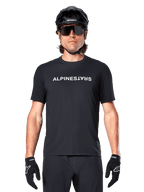 Cycliste de VTT portant le maillot à manches courtes Alpinestars A-Dura Switch noir, avec logo textuel blanc sur la poitrine, casque noir, grandes lunettes de soleil et gants noirs, équipement de cyclisme professionnel pour le trail