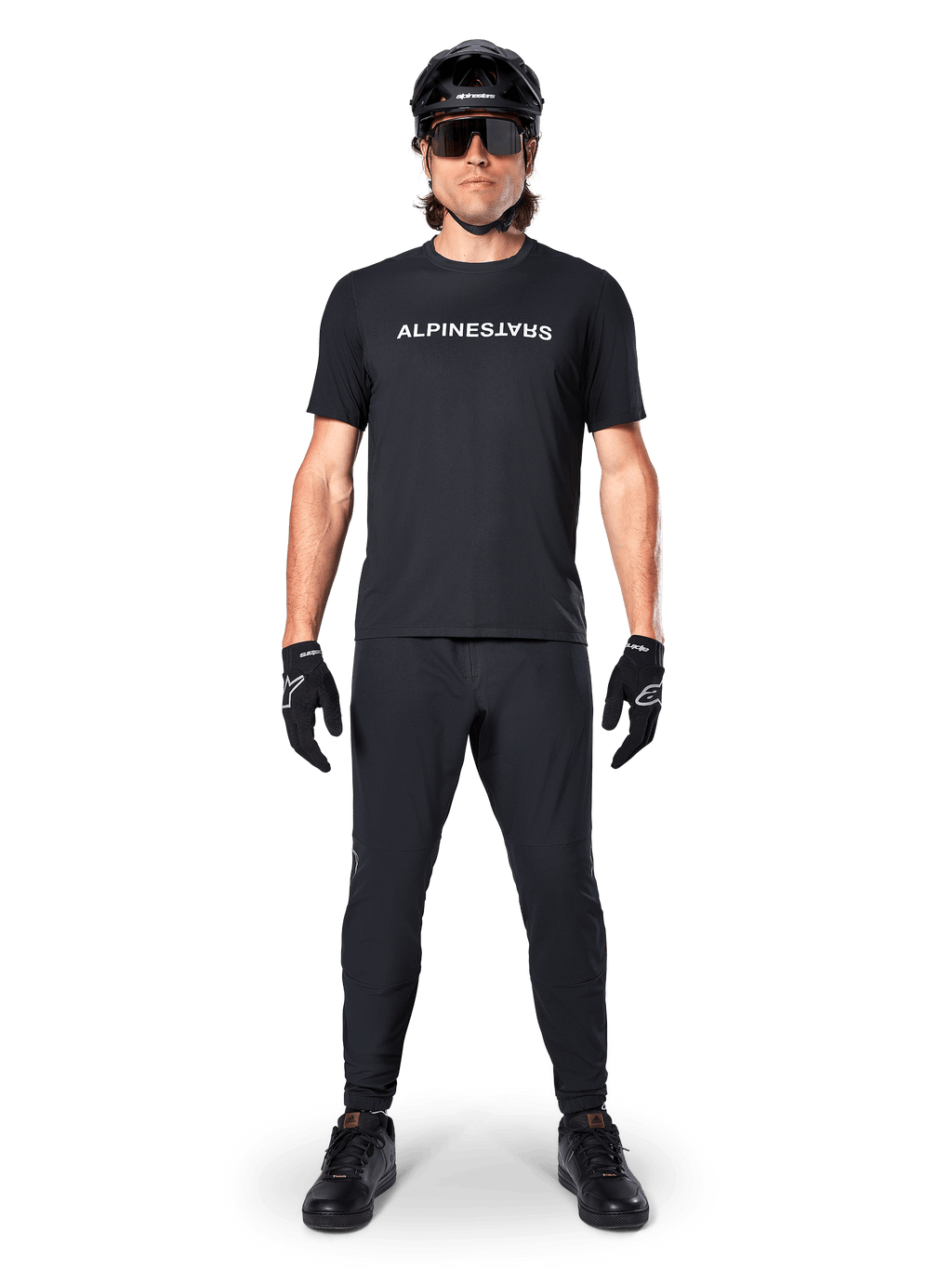 Cycliste de VTT portant le maillot à manches courtes Alpinestars A-Dura Switch en noir, avec un logo blanc stylisé sur la poitrine, associé à un pantalon de VTT, un casque et des gants noirs assortis, tenue de cyclisme complète prête pour les sentiers
