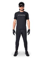 Cycliste de VTT portant le maillot à manches courtes Alpinestars A-Dura Switch en noir, avec un logo blanc stylisé sur la poitrine, associé à un pantalon de VTT, un casque et des gants noirs assortis, tenue de cyclisme complète prête pour les sentiers