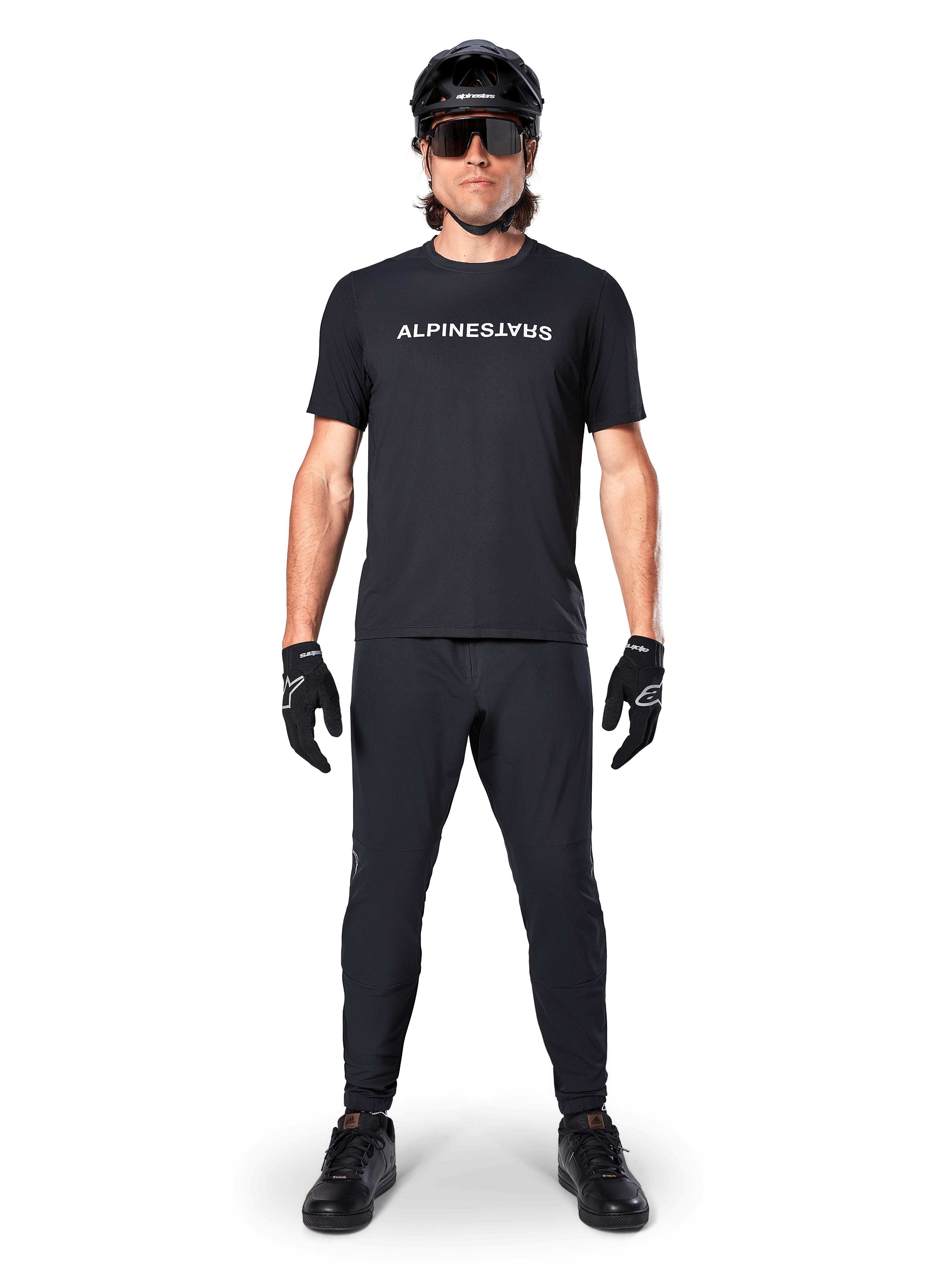 Cycliste de VTT portant le maillot à manches courtes Alpinestars A-Dura Switch en noir, avec un logo blanc stylisé sur la poitrine, associé à un pantalon de VTT, un casque et des gants noirs assortis, tenue de cyclisme complète prête pour les sentiers