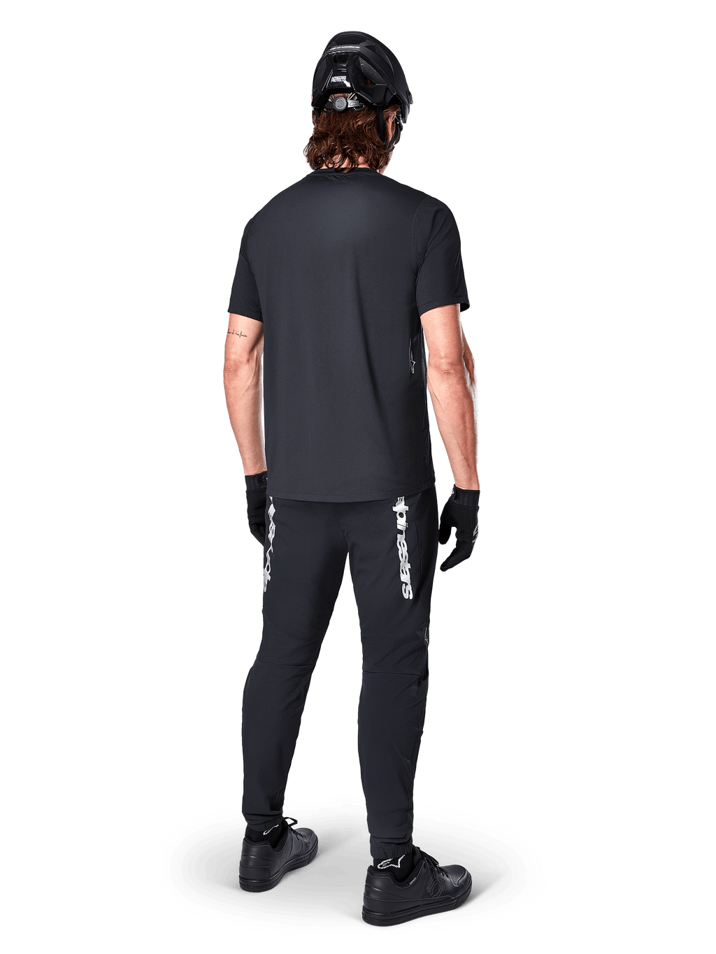 Maillot à manches courtes Alpinestars A-Dura Switch, noir, vue de dos d'un cycliste portant une tenue de VTT complète, incluant un pantalon noir assorti avec lettrage blanc, un casque noir et des gants, équipement de cyclisme de performance pour le trail