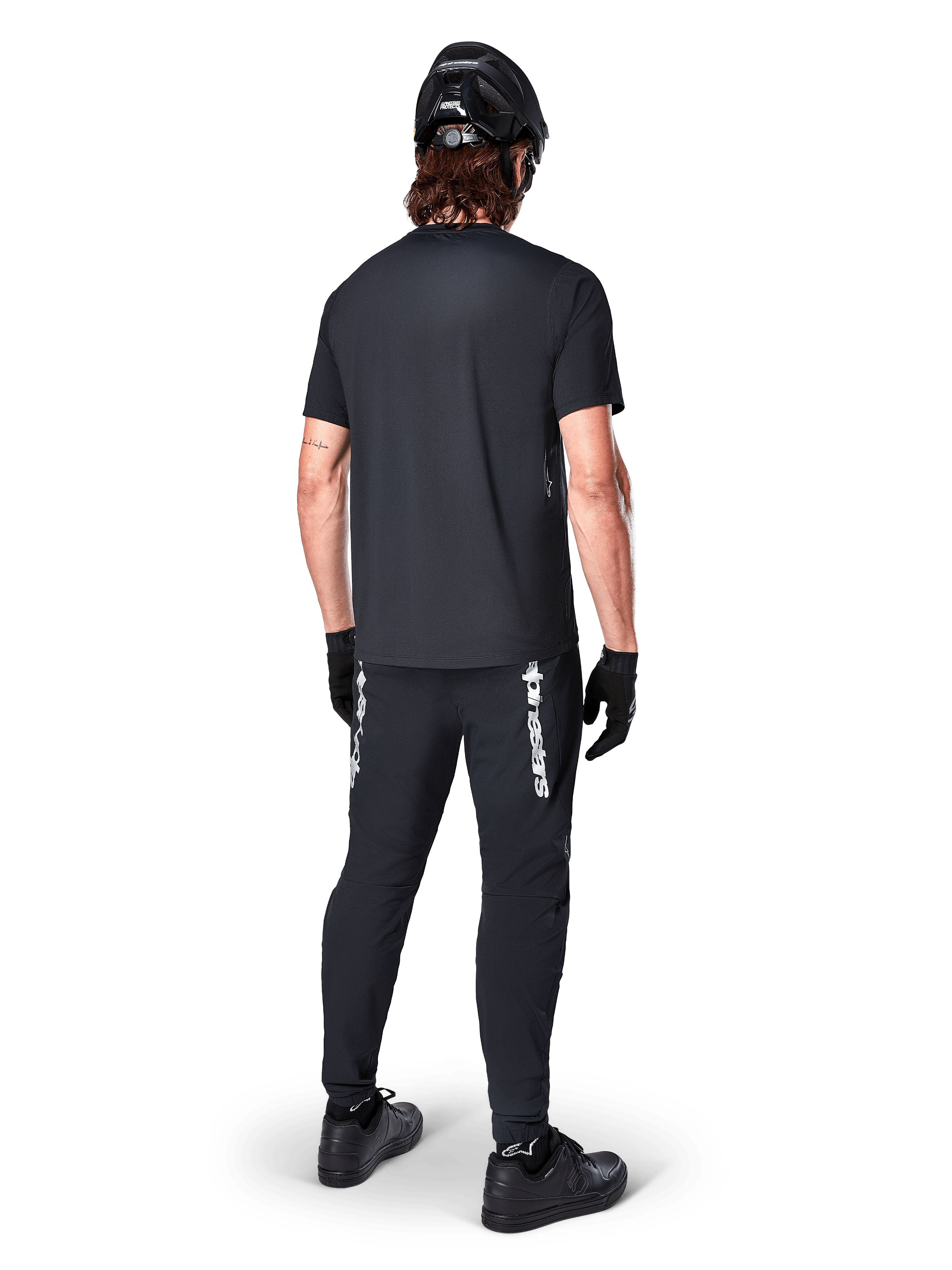 Maillot à manches courtes Alpinestars A-Dura Switch, noir, vue de dos d'un cycliste portant une tenue de VTT complète, incluant un pantalon noir assorti avec lettrage blanc, un casque noir et des gants, équipement de cyclisme de performance pour le trail