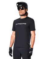 Maillot à manches courtes Alpinestars A-Dura Switch, noir, arborant un logo textuel blanc sur la poitrine, porté par un cycliste muni d'un casque de VTT noir et de lunettes de soleil, vêtement technique conçu pour le trail