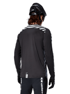 Maillot à manches longues Alpinestars A-Dura Dri Oscar, Noir réfléchissant, vue arrière montrant des rayures diagonales réfléchissantes sur les épaules et un pan arrière plus long, porté avec un casque et des gants de VTT noirs, équipement technique pour le VTT