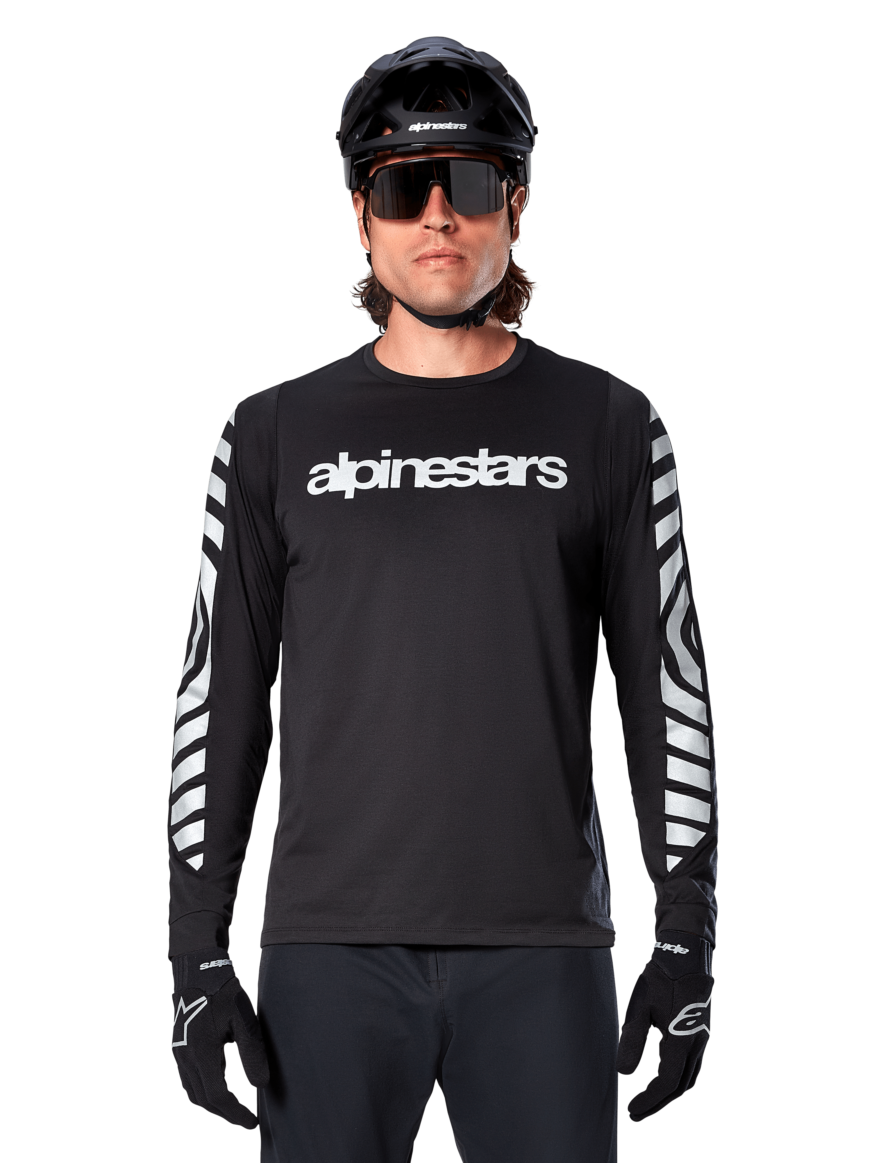 Cycliste portant le maillot à manches longues Alpinestars A-Dura Dri Oscar, Noir Réfléchissant, avec lettrage Alpinestars blanc sur la poitrine et graphismes argentés réfléchissants sur les manches, accompagné d'un casque de VTT noir, de lunettes de soleil et de gants noirs, tenue de cyclisme haute performance