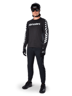 Maillot à manches longues Alpinestars A-Dura Dri Oscar, noir réfléchissant, arborant un lettrage Alpinestars blanc sur la poitrine et des motifs graphiques réfléchissants sur les manches, porté avec un pantalon de VTT noir, un casque et des gants, équipement technique de VTT